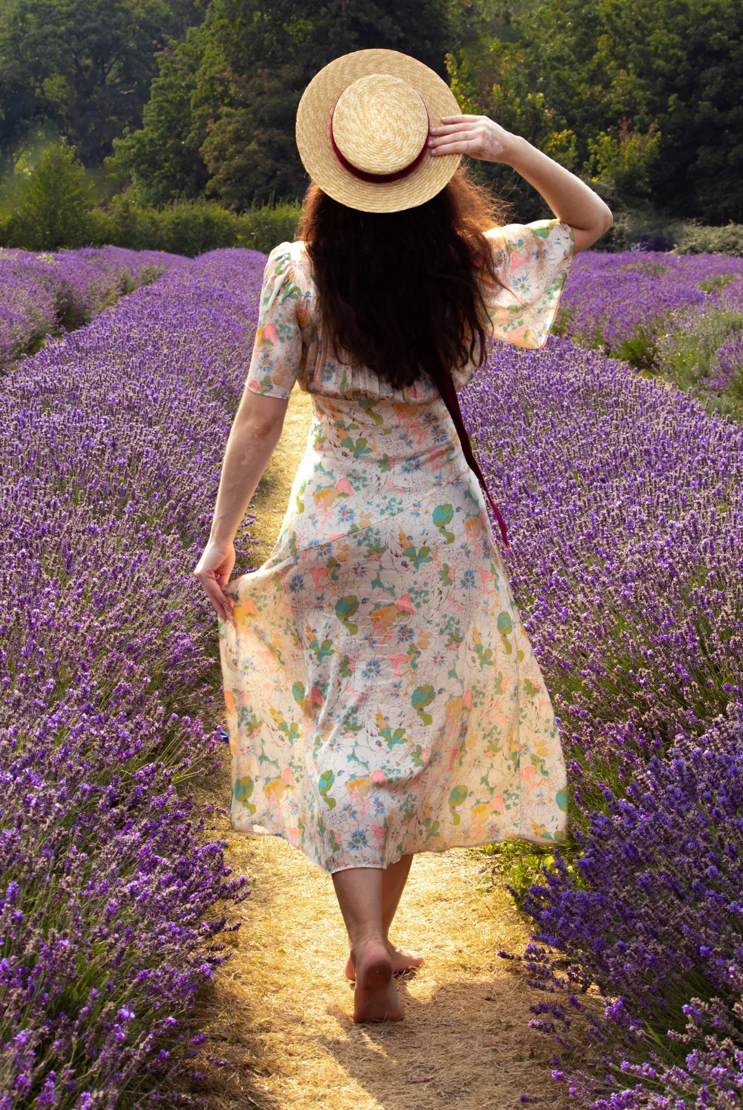 A woman walking barefoot through a lavender field, wearing a floral dress and a straw hat. She holds her dress with one hand while adjusting her hat with the other.