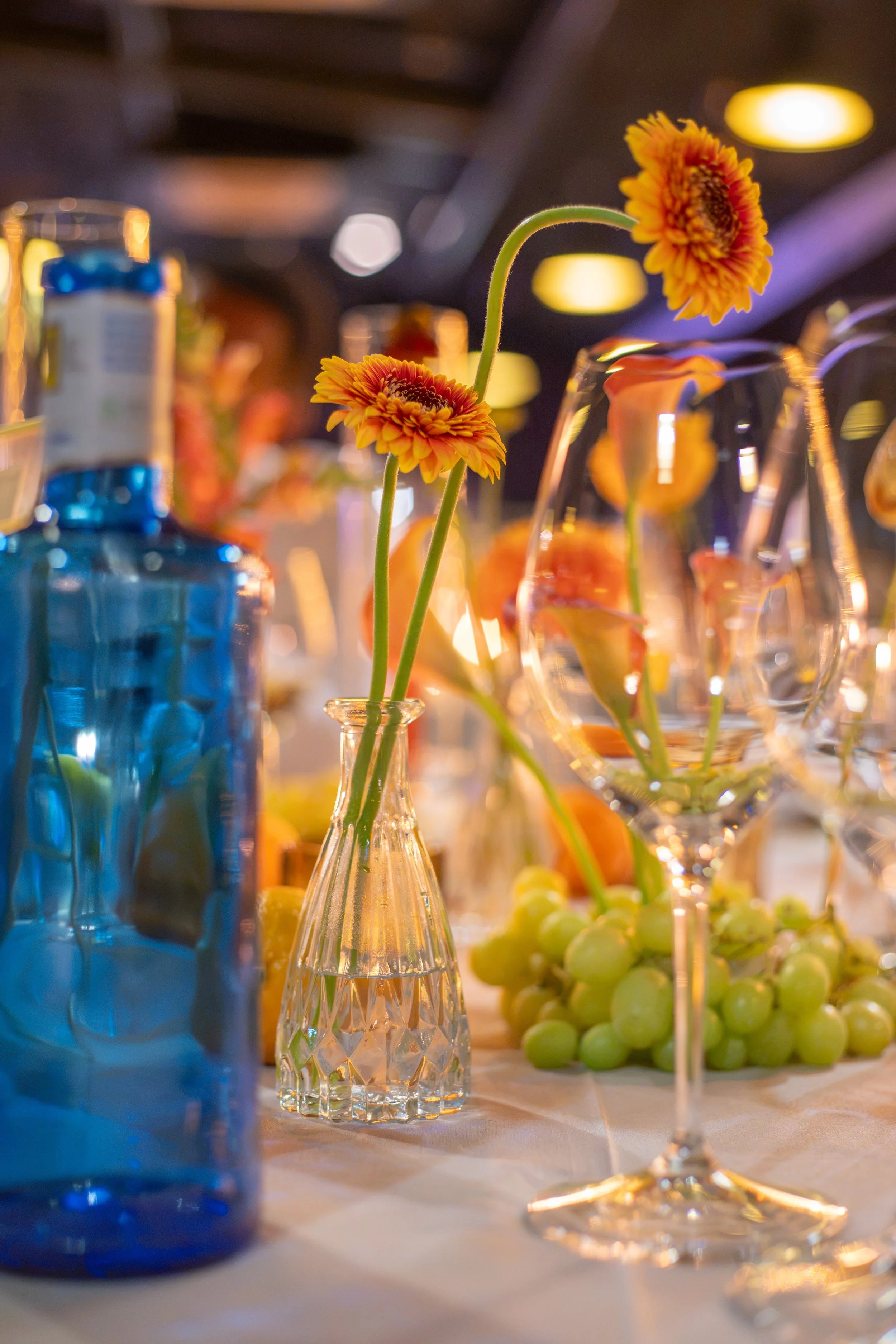 Close-up of a flower arrangement with yellow-orange flowers in a clear glass vase, surrounded by colorful bottles and glasses on a table, with warm lighting and a blurred background.