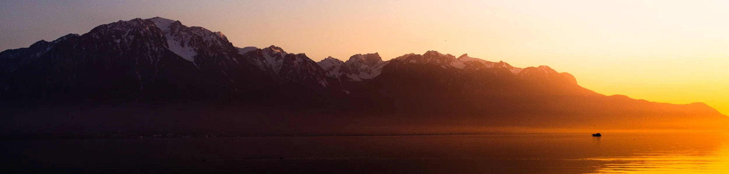 Sunset view over lake Geneva showing a mountain range with snow-capped peaks across the lake, and a small boat on the water.