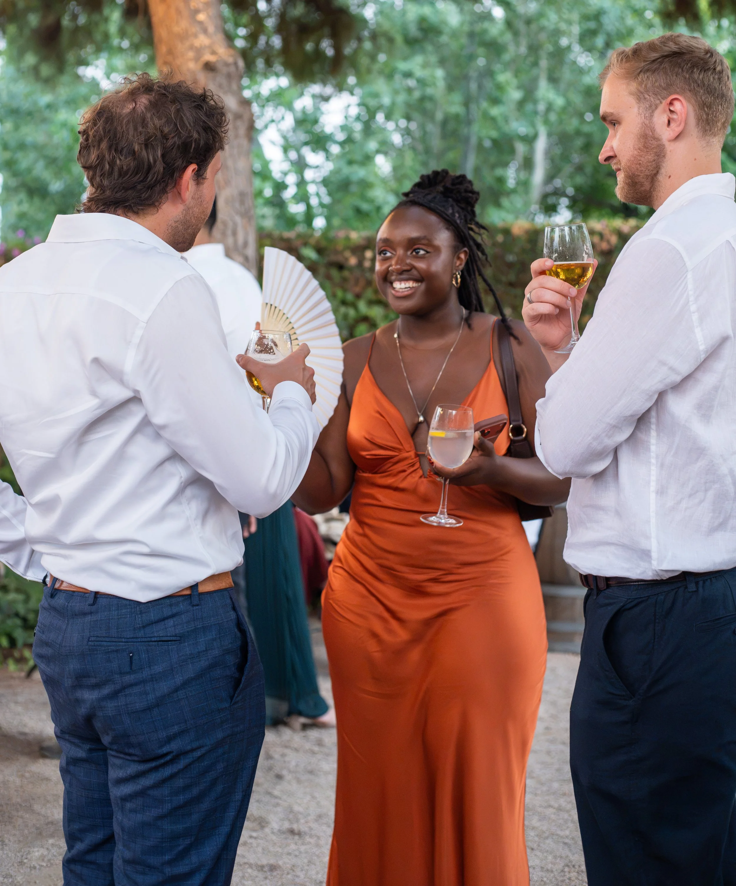 Three people socialising outdoors at a wedding reception, two white men in white shirts and a black woman in an orange dress, holding drinks.