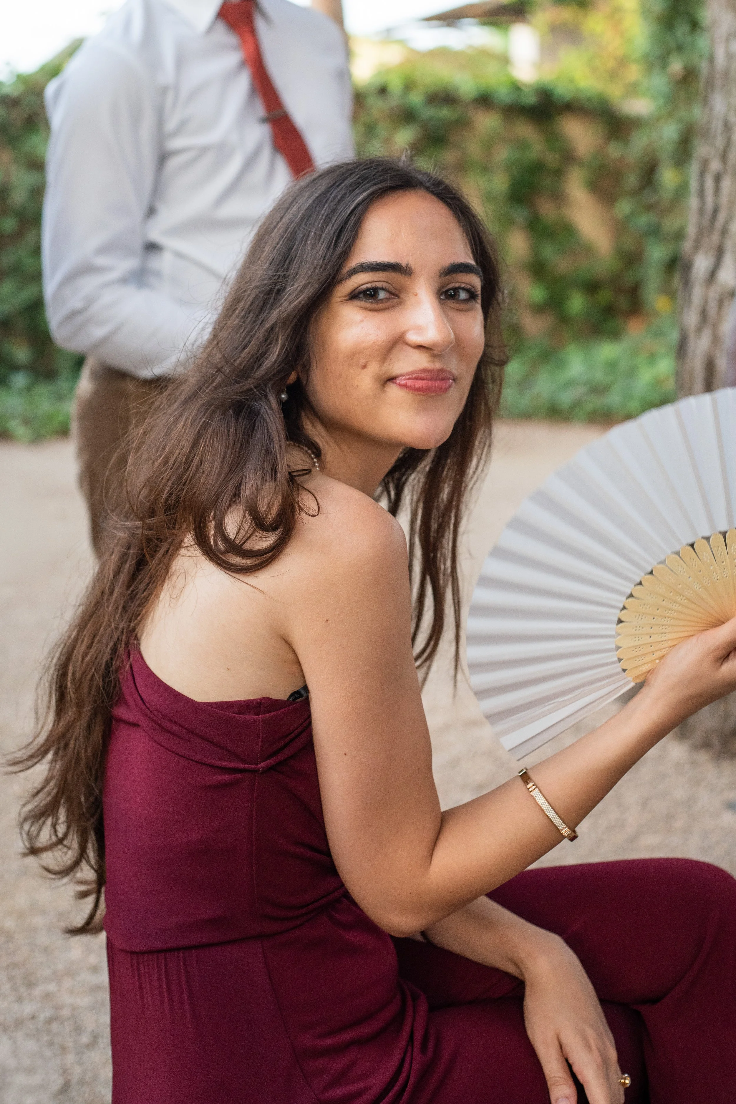 A woman with long brown hair sitting outdoors at a wedding reception, wearing a burgundy dress, holding a decorative white and beige fan, and smiling at the camera. 