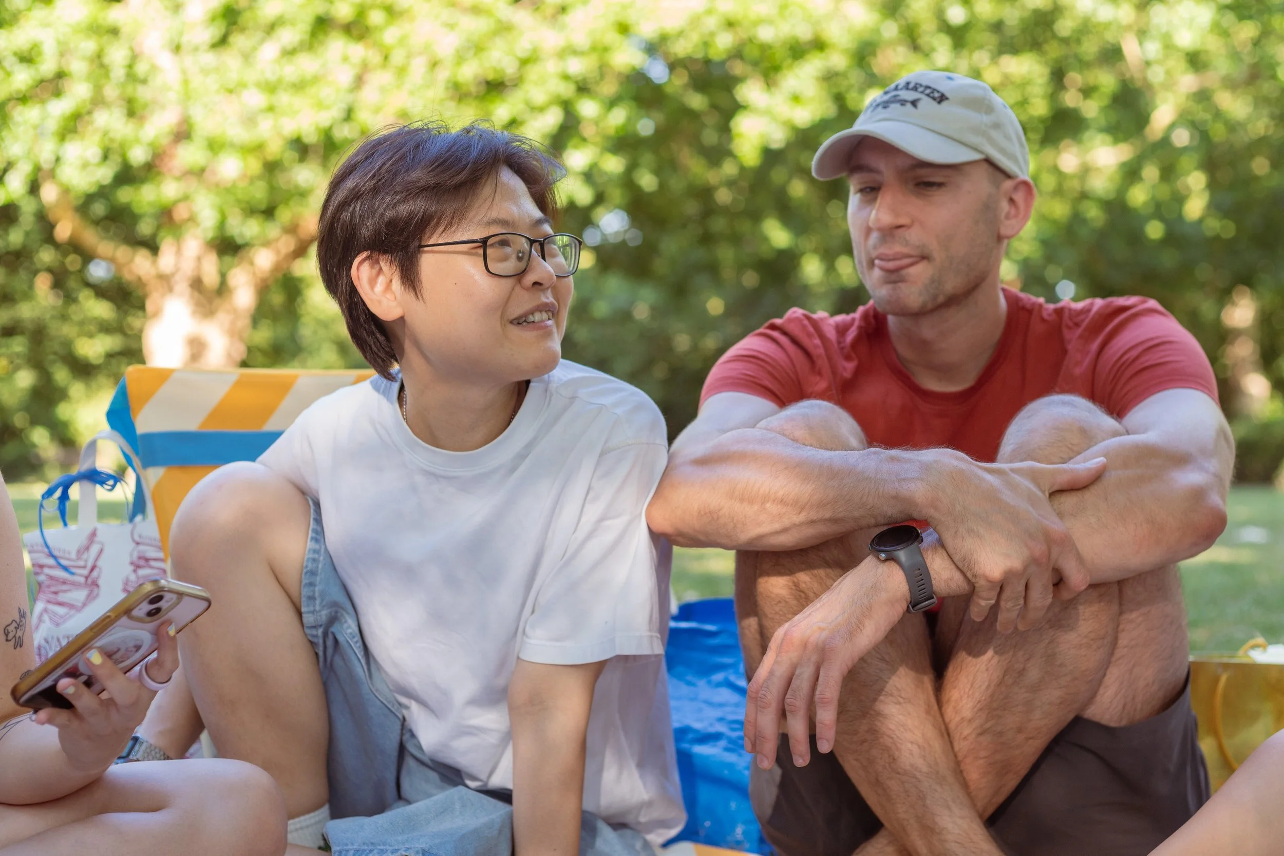 Two people, a woman with short brown hair and glasses and a man wearing a white cap, are sitting close together outdoors among green trees, engaging in conversation with relaxed expressions. The woman is holding a phone in her right hand, and the man