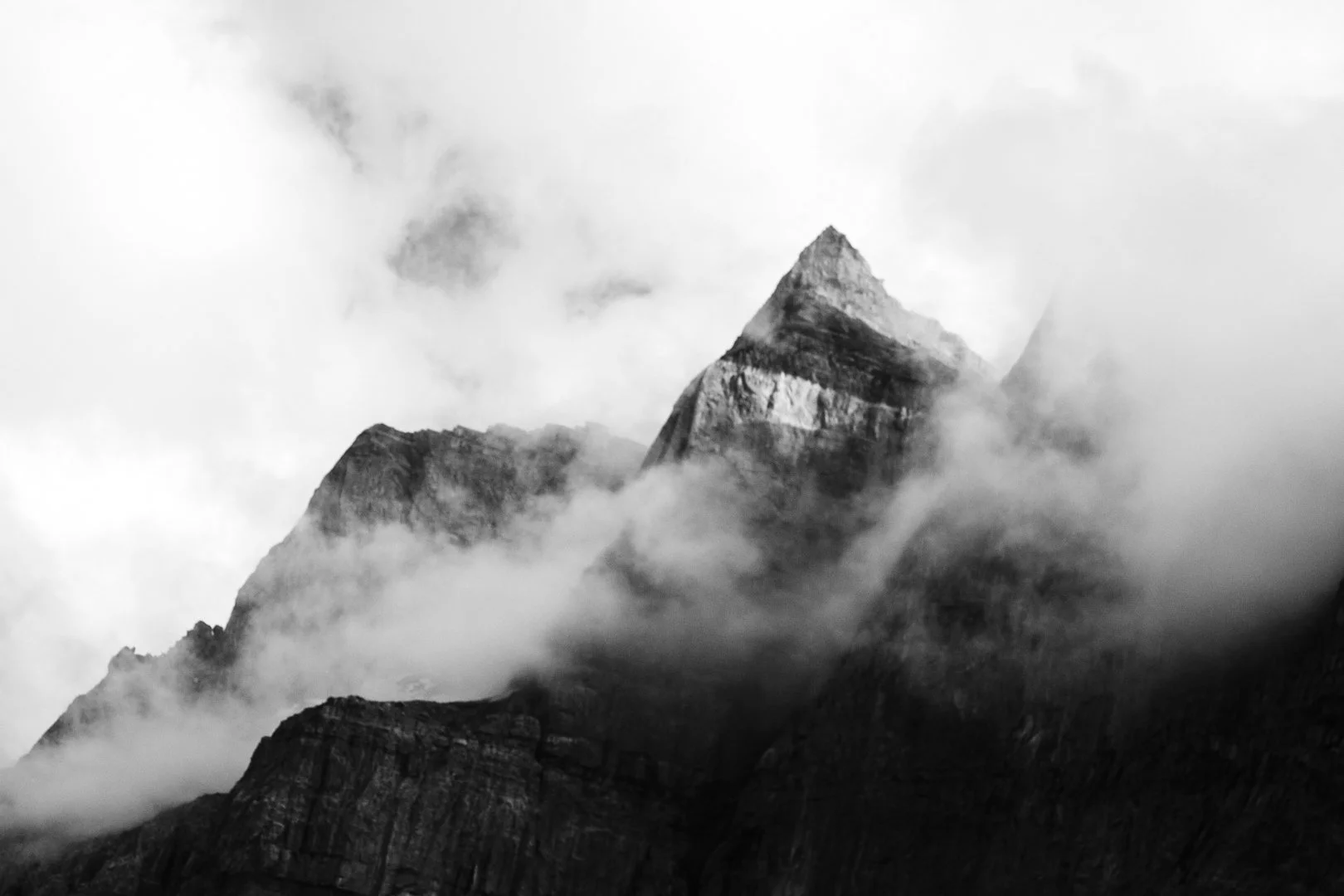 Black and white photograph of a tall, jagged mountain peak partially obscured by clouds and mist.