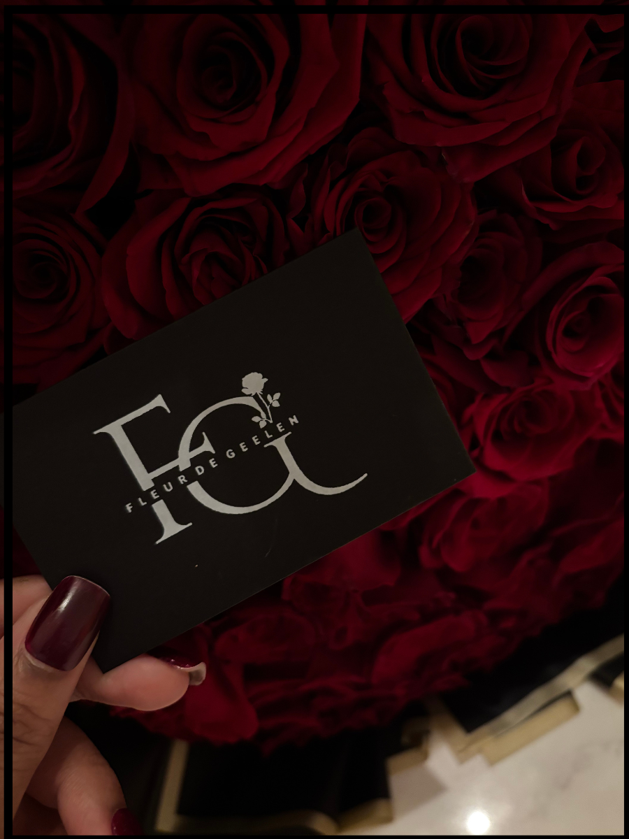 A hand holding a black card with the logo and text 'Fleur de Gelin' in front of a large bouquet of red roses.