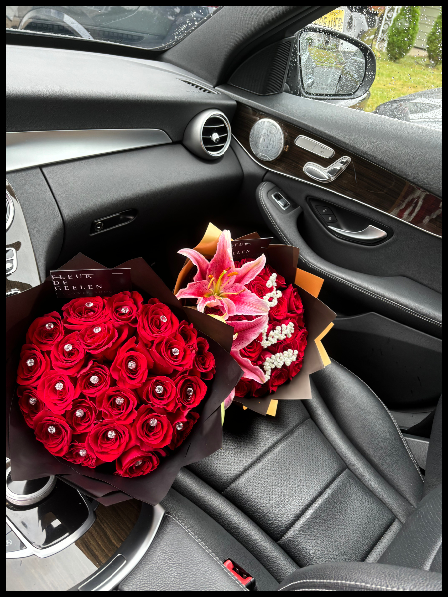 Inside a car, two bouquets of red and pink flowers are placed on the passenger seat. The bouquets include roses, lilies, and small white flowers. The car's interior features black seats, a dashboard with wood trim, and a side mirror with a rainy window.