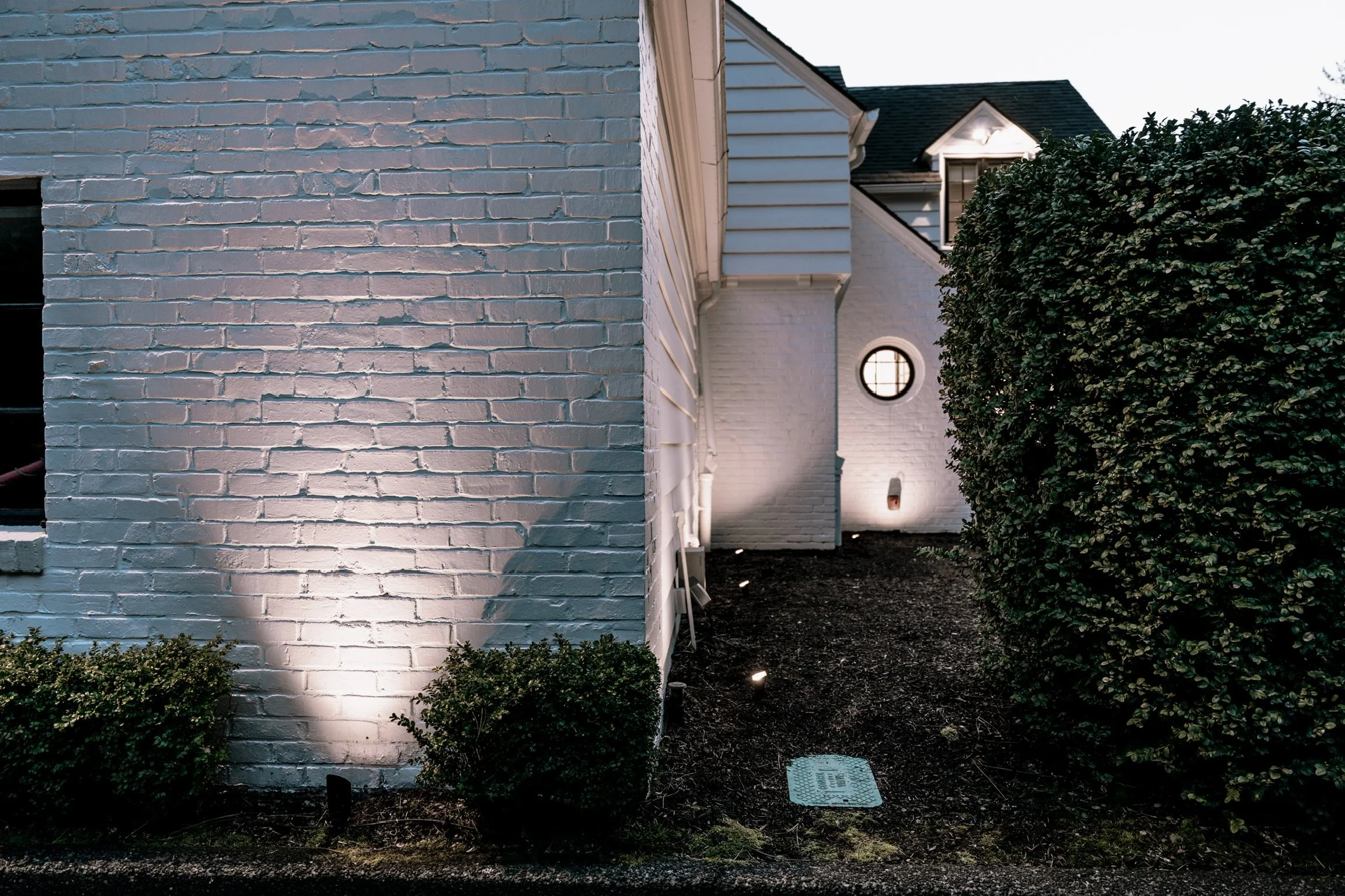 Nighttime view of the exterior of a white house with a brick wall, bush, and pathway lit by ground lights.