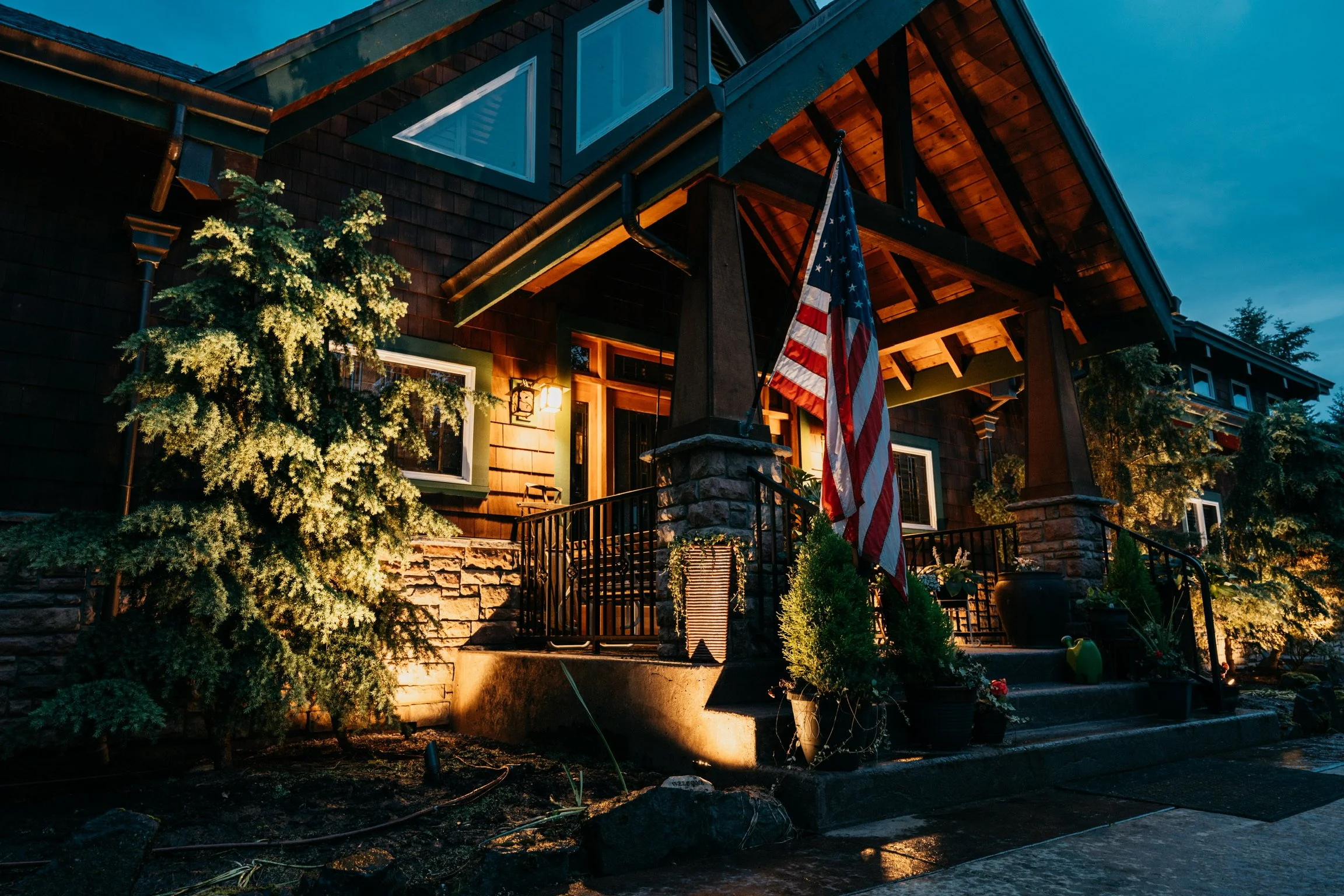 A house at dusk with exterior lighting, American flag on the porch, surrounded by trees and plants.