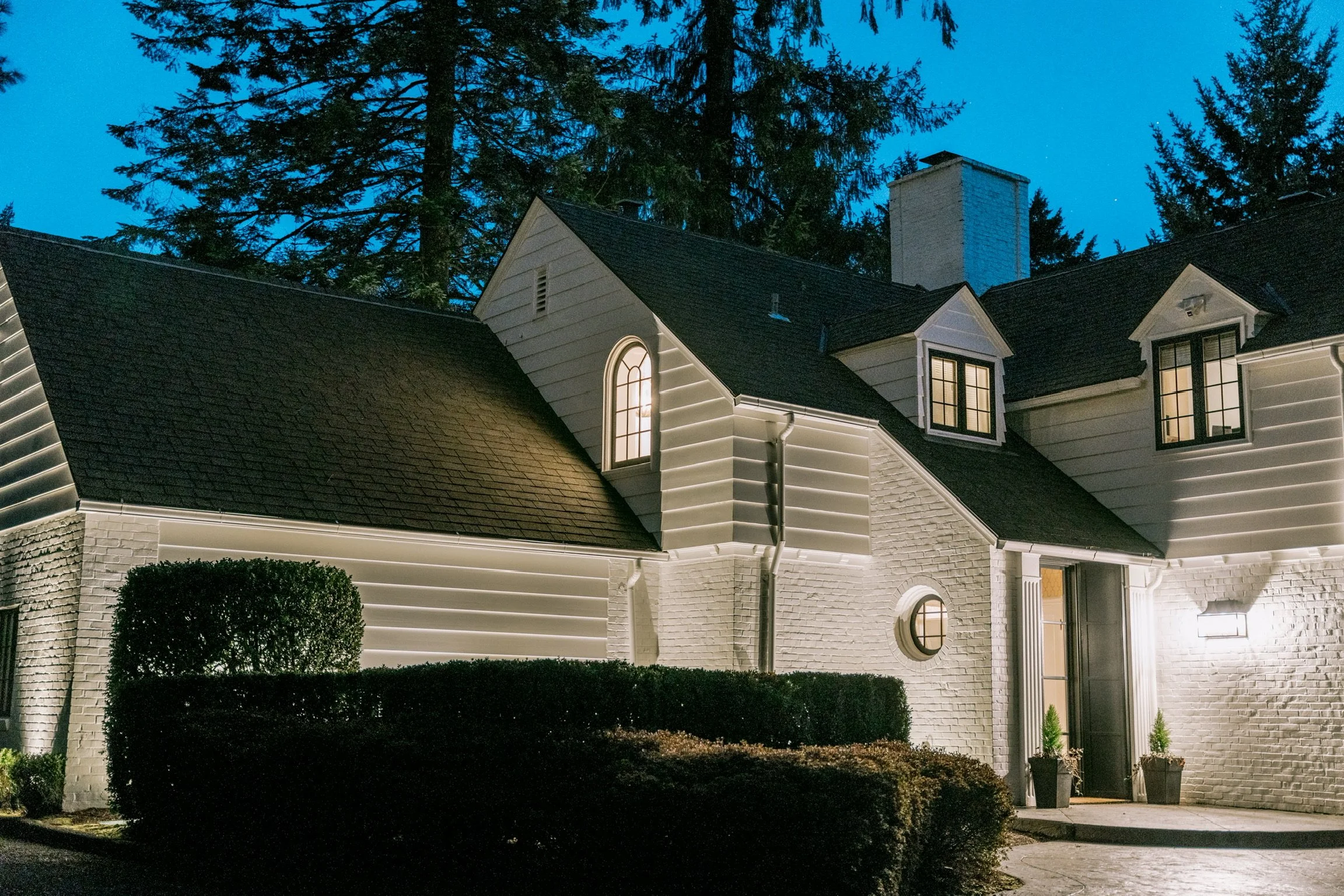 A night view of a house with exterior lighting, white brick walls, black roof, and illuminated windows, with trees in the background.