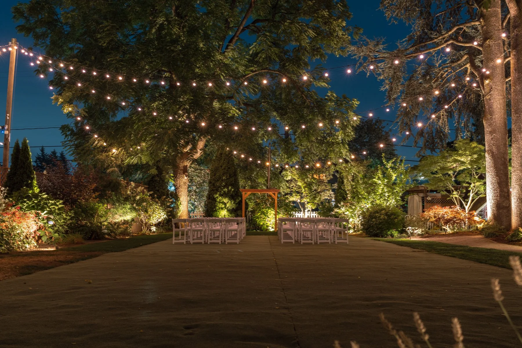 Night outdoor wedding setup with string lights hanging over chairs arranged in rows, surrounded by trees and bushes with ambient lighting, on a paved pathway.