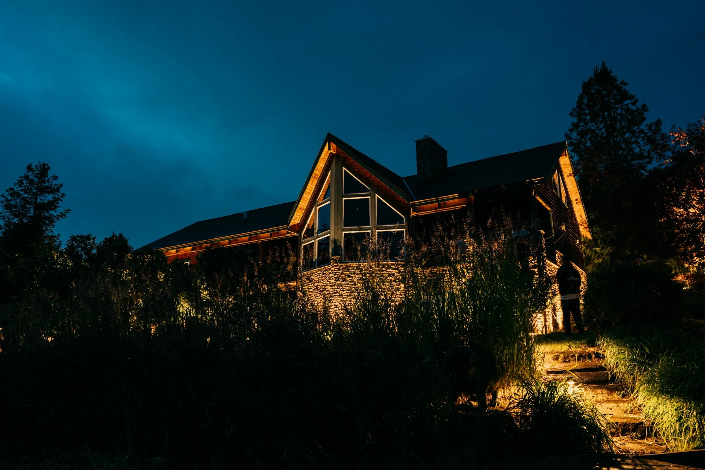 A modern house illuminated at night, with large windows and a stone facade, surrounded by trees and tall grass, with a person standing near the steps.