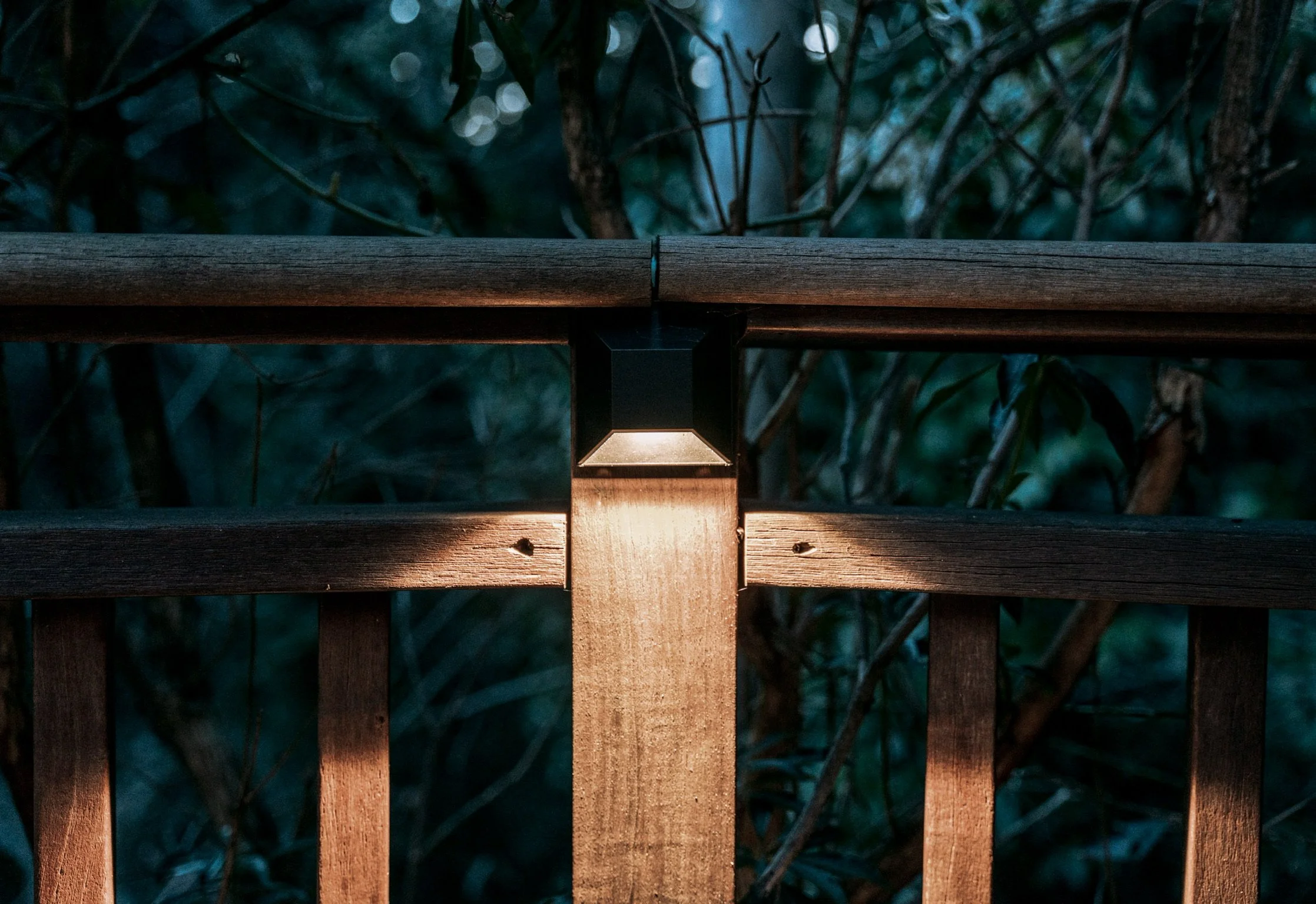 Close-up of a wooden fence with a black solar-powered light fixture attached, illuminating the wood during dusk or nighttime.