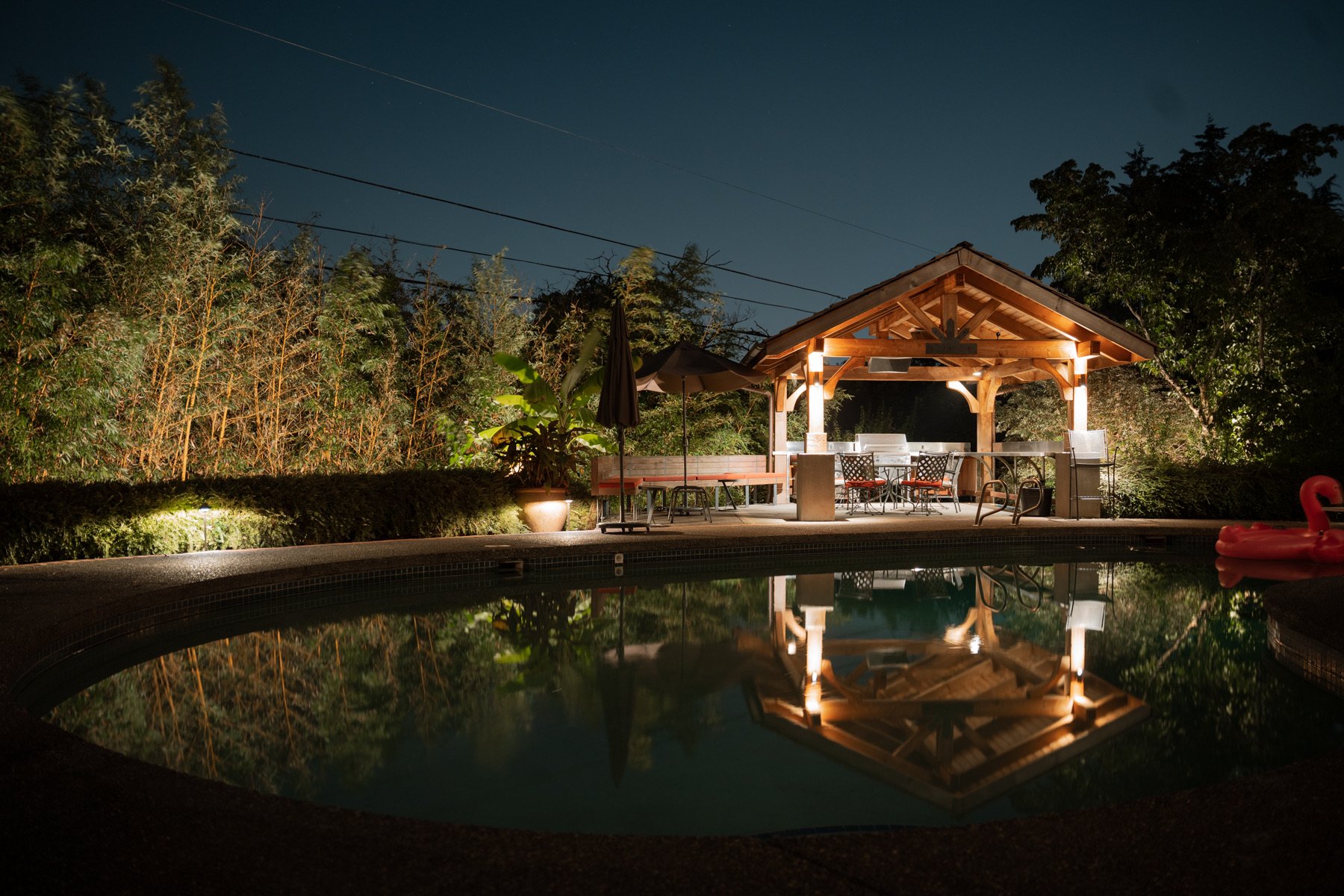 Nighttime scene of a backyard pool with a illuminated gazebo, outdoor furniture, and lush trees and bushes in the background, with their reflection visible in the pool.