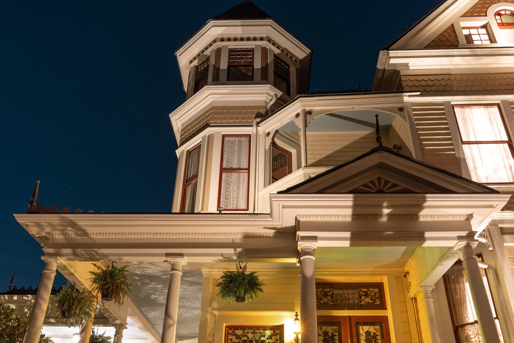A historic Victorian-style house illuminated at night, showing detailed architecture with a prominent tower, decorative trim, hanging plants, and warm outdoor lighting.