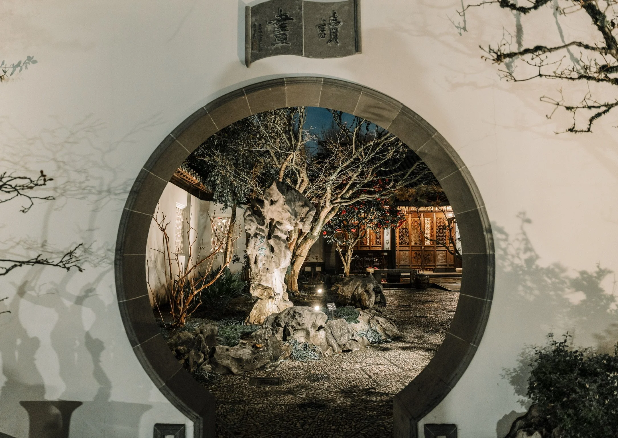 A traditional Chinese garden scene viewed through a circular moon gate. It features a rock formation, trees with twisting branches, and a wooden building in the background, illuminated at night.