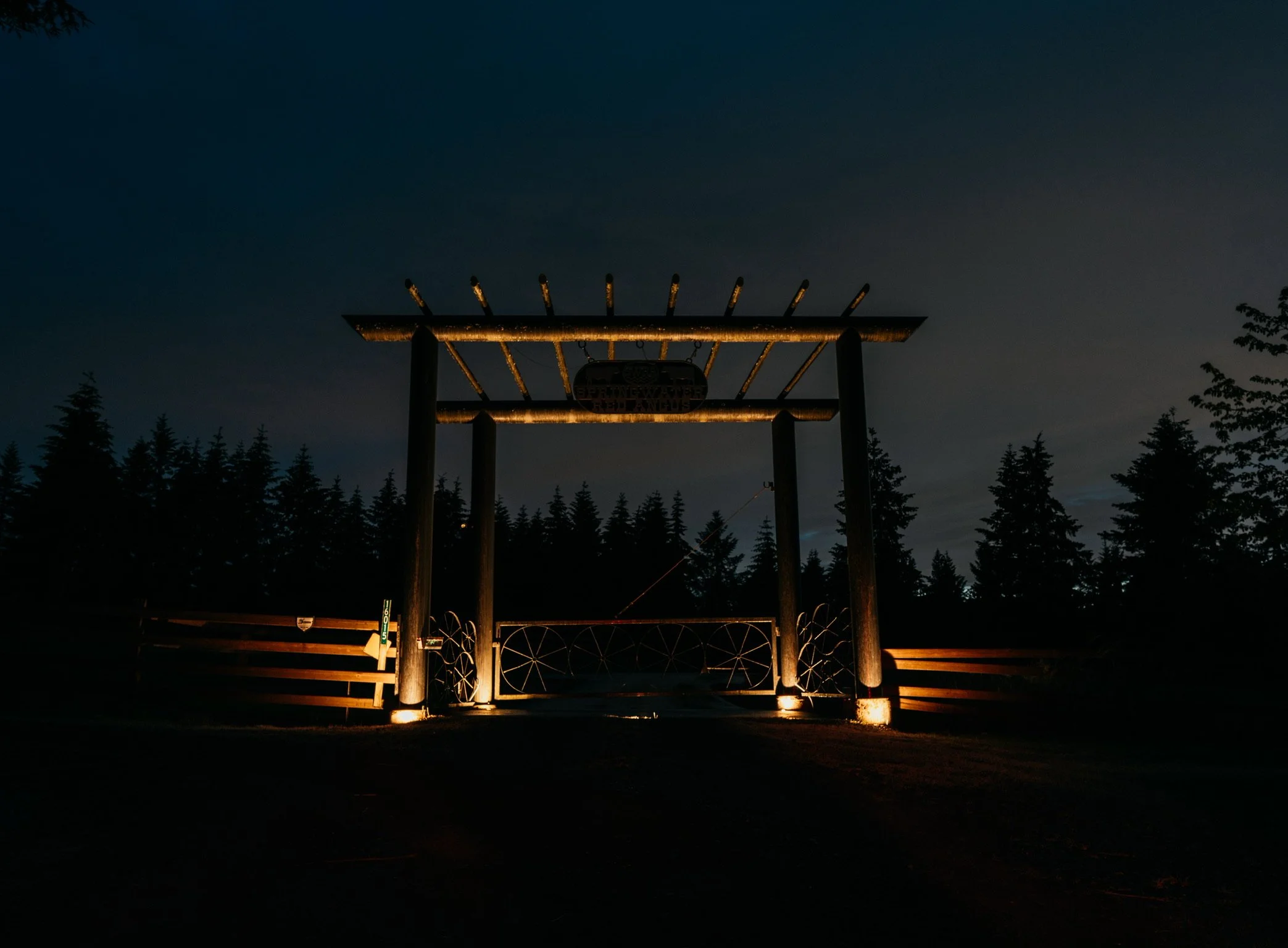 Dark night scene showing the illuminated entrance gate of a park or reserve, with a sign hanging above, surrounded by silhouettes of trees.