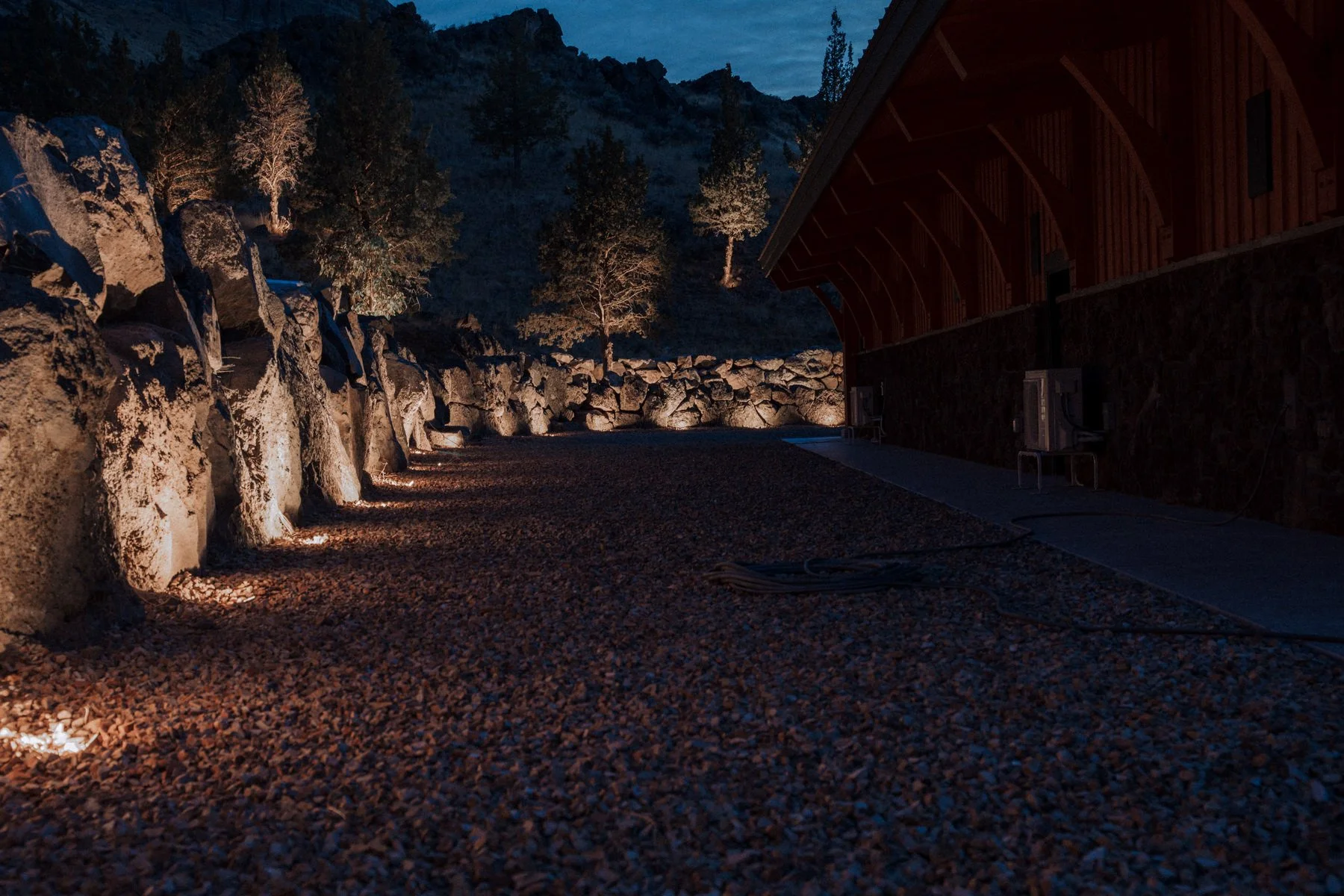 Nighttime exterior view of a building with a gravel pathway, lit rocks, and trees in the background.