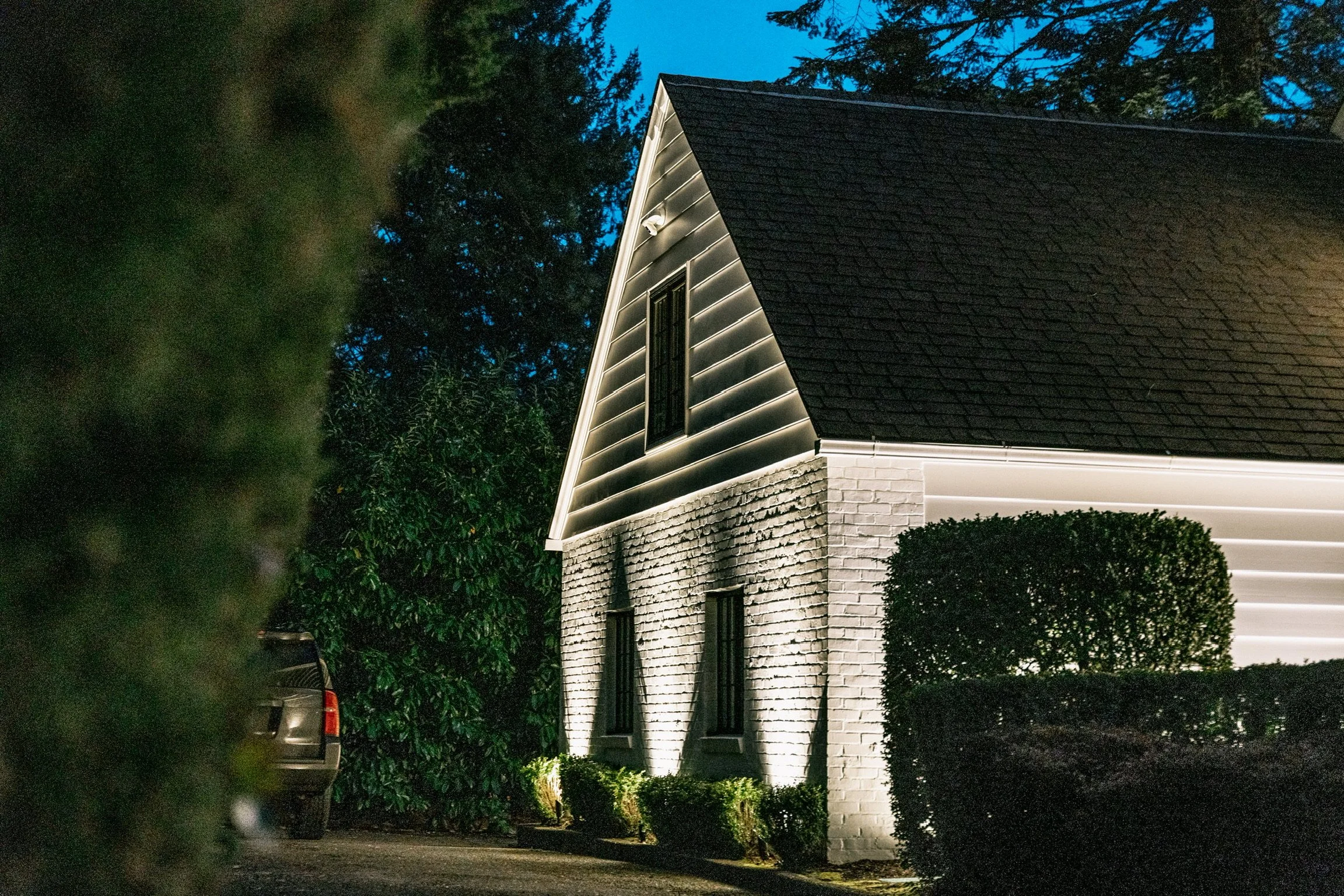 Exterior of a house illuminated at night with bright white lights highlighting the brick and siding walls, a window, and well-trimmed bushes in front.
