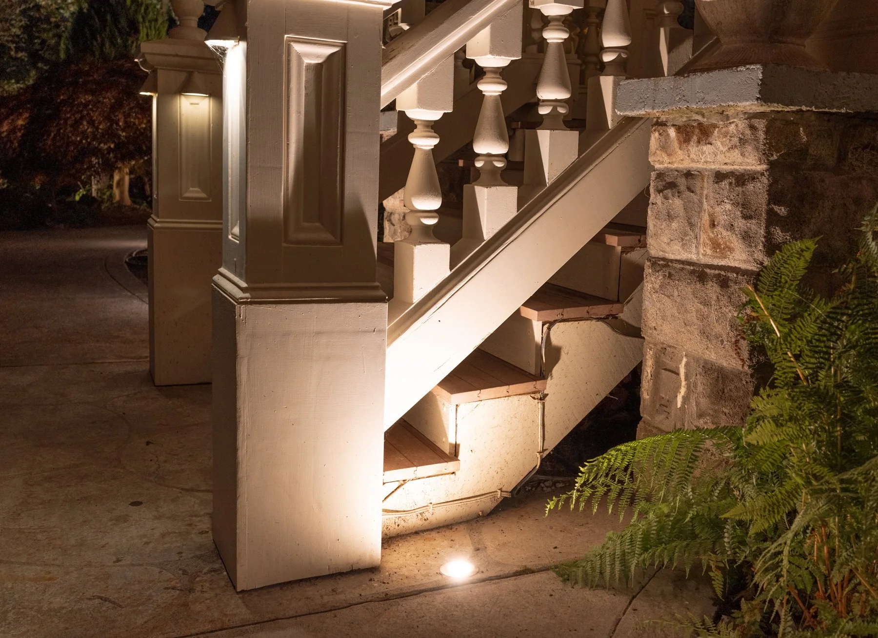Close-up of a staircase with painted white wood balusters and newel post, and a brick wall on the right, illuminated by a ground light at night.