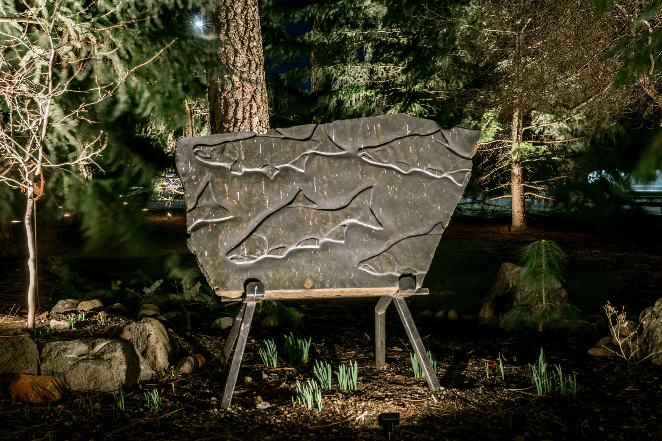 Outdoor art installation featuring a large, irregularly shaped dark stone slab with carved abstract lines, mounted on a metal stand, surrounded by trees and small plants at night.