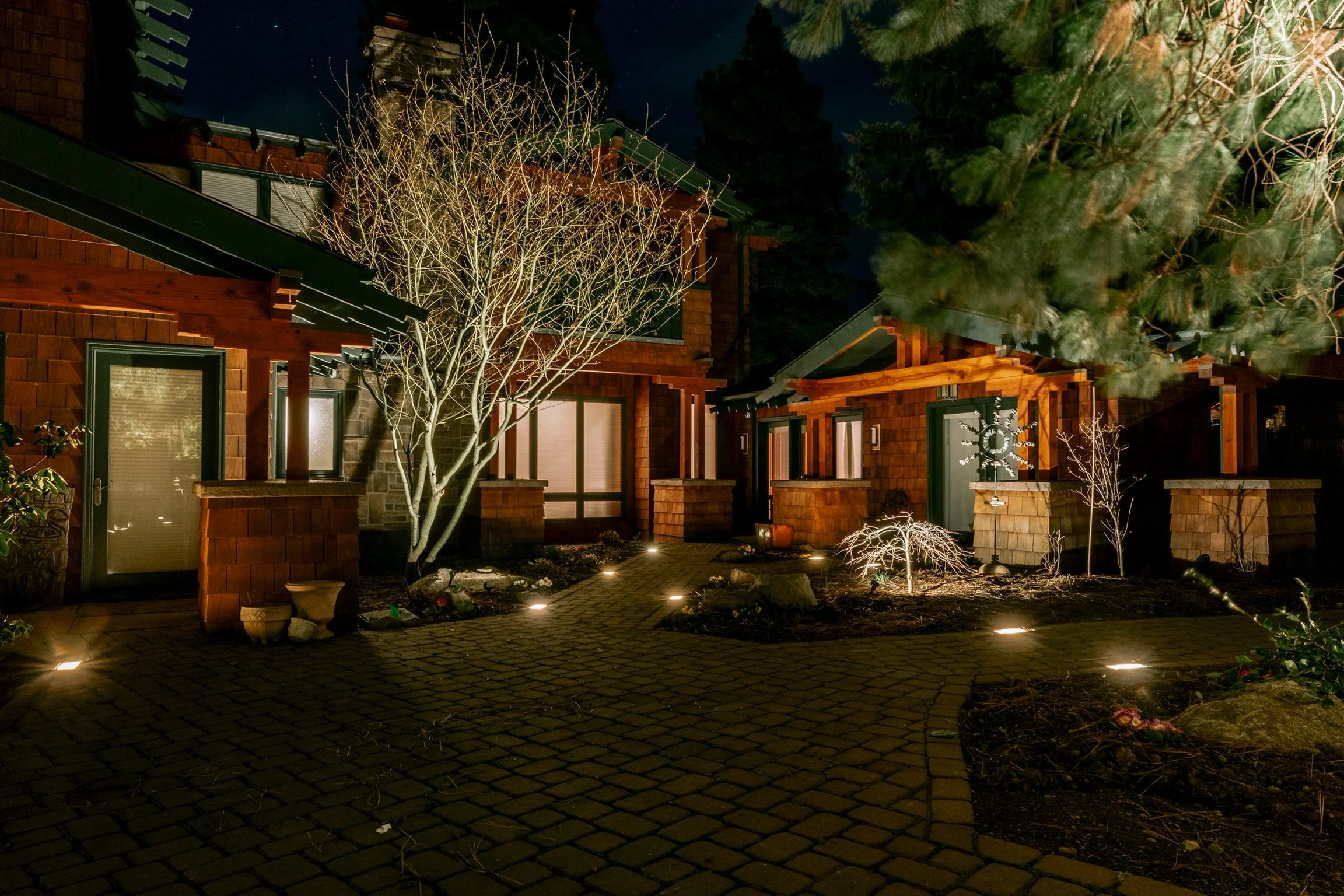 Nighttime view of a residential courtyard illuminated with outdoor lighting. Stone-paved path, leafless trees, shrubbery, and wooden houses with large windows.