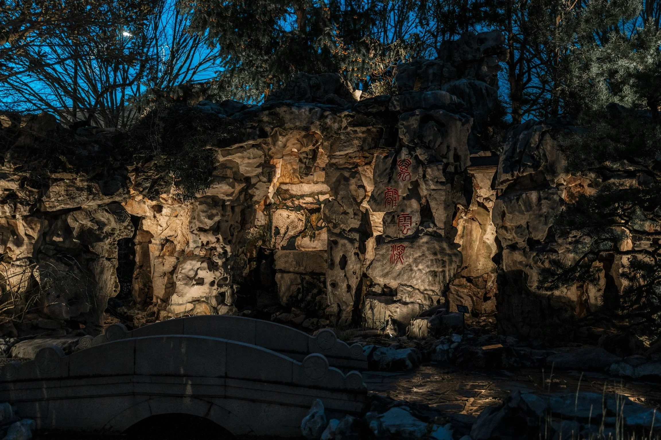 Night view of a rock garden with illuminated rocks and Japanese inscriptions, surrounded by trees.