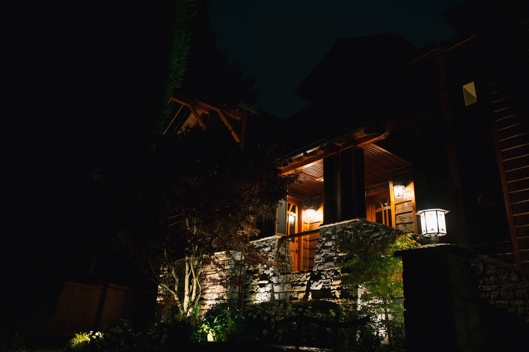 A nighttime view of a modern house with warm exterior lights illuminating the entrance, stone stairs, wooden accents, and landscaping with trees and plants.