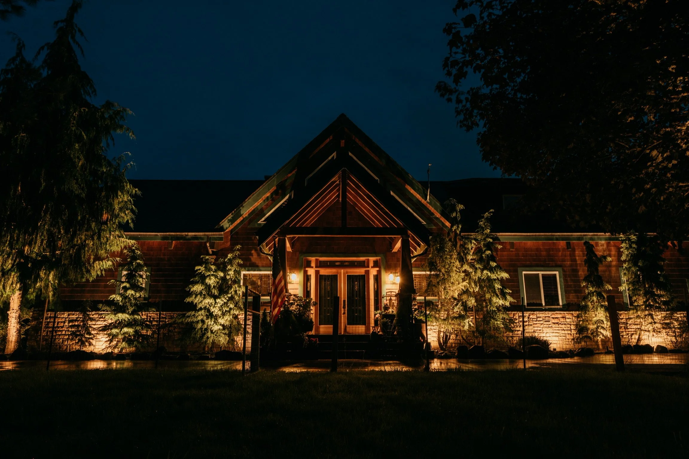 Night view of a house with illuminated front porch, surrounded by trees and a fence.