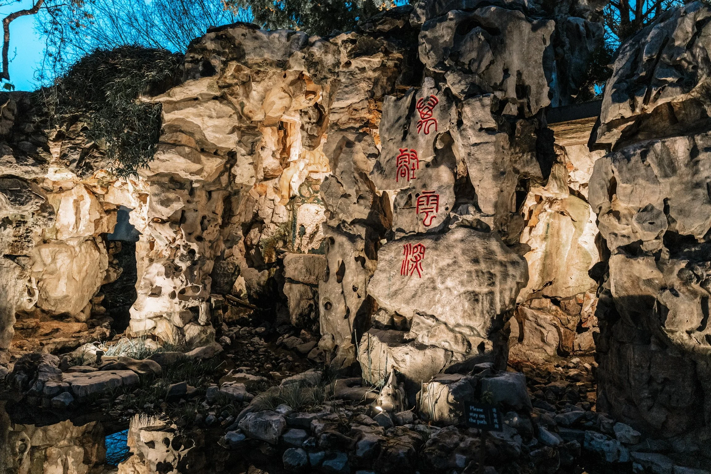 Nighttime scene of a stone formation with red Chinese characters inscribed on large rocks, illuminated by soft lighting, with reflections in a nearby water pool, and a small sign at the bottom reading 'Please stay on the path.'