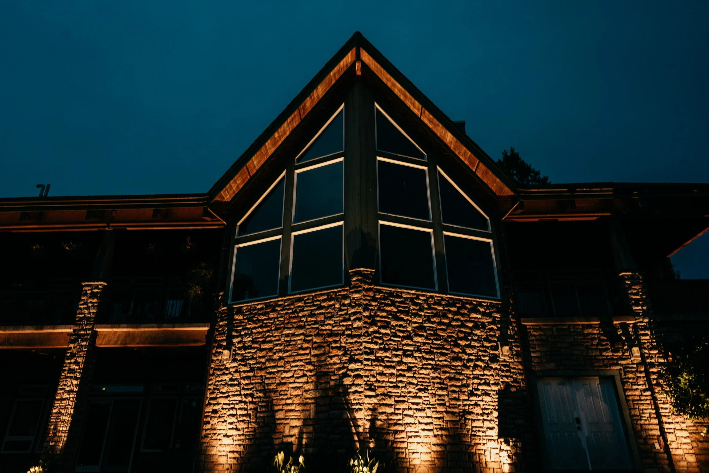 A modern house at night with illuminated exterior lights highlighting the brick and large glass windows.