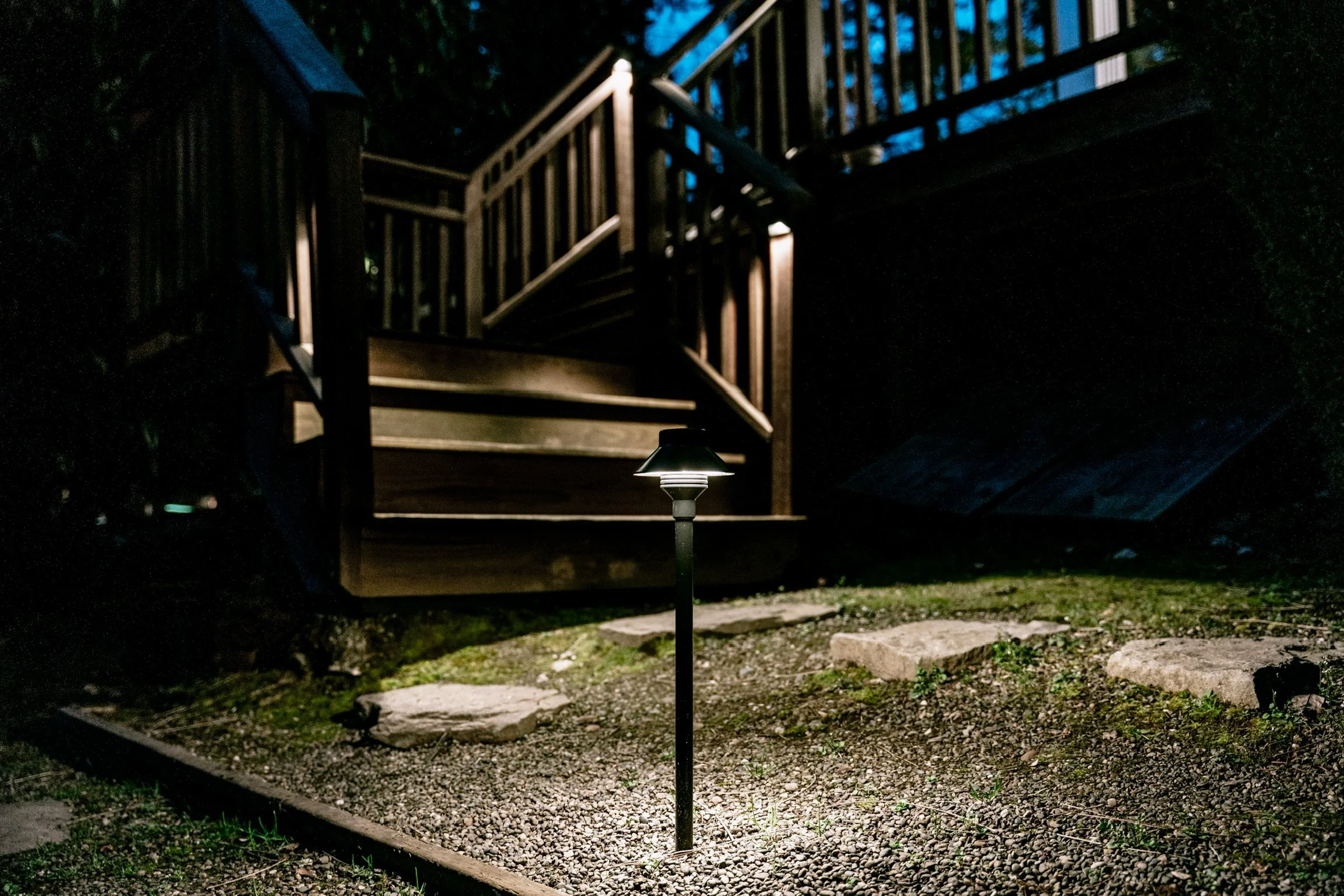 Nighttime outdoor scene with a lit pathway light in the foreground, a gravel path, and a wooden staircase leading to a deck or porch in the background.