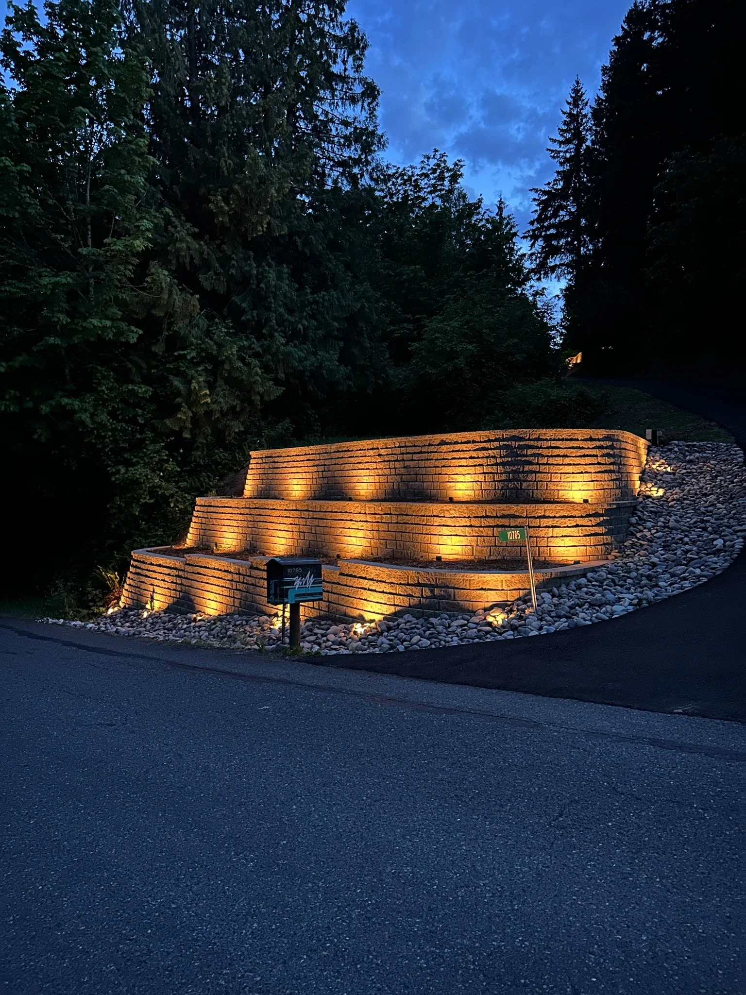A brick retaining wall illuminated with warm lights at night, with a mailbox in front and trees in the background.