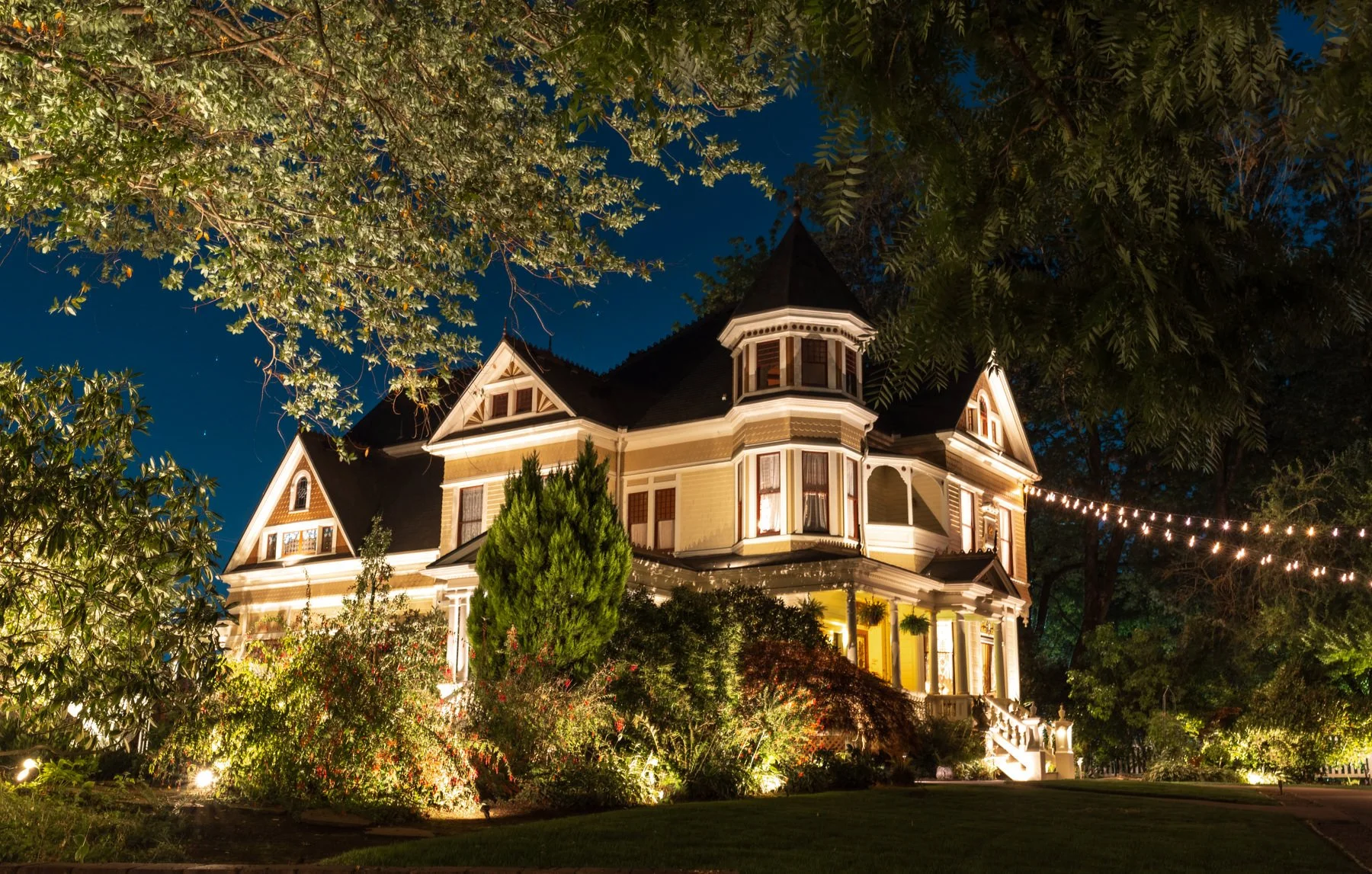 A large, historic Victorian house illuminated at night, with string lights across the garden and surrounded by trees and bushes.