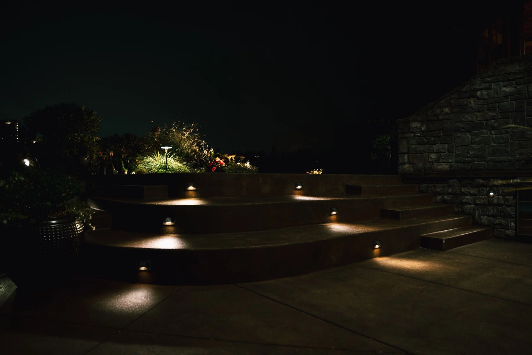 Nighttime outdoor scene with steps illuminated by small ground lights, garden plants, and a brick wall