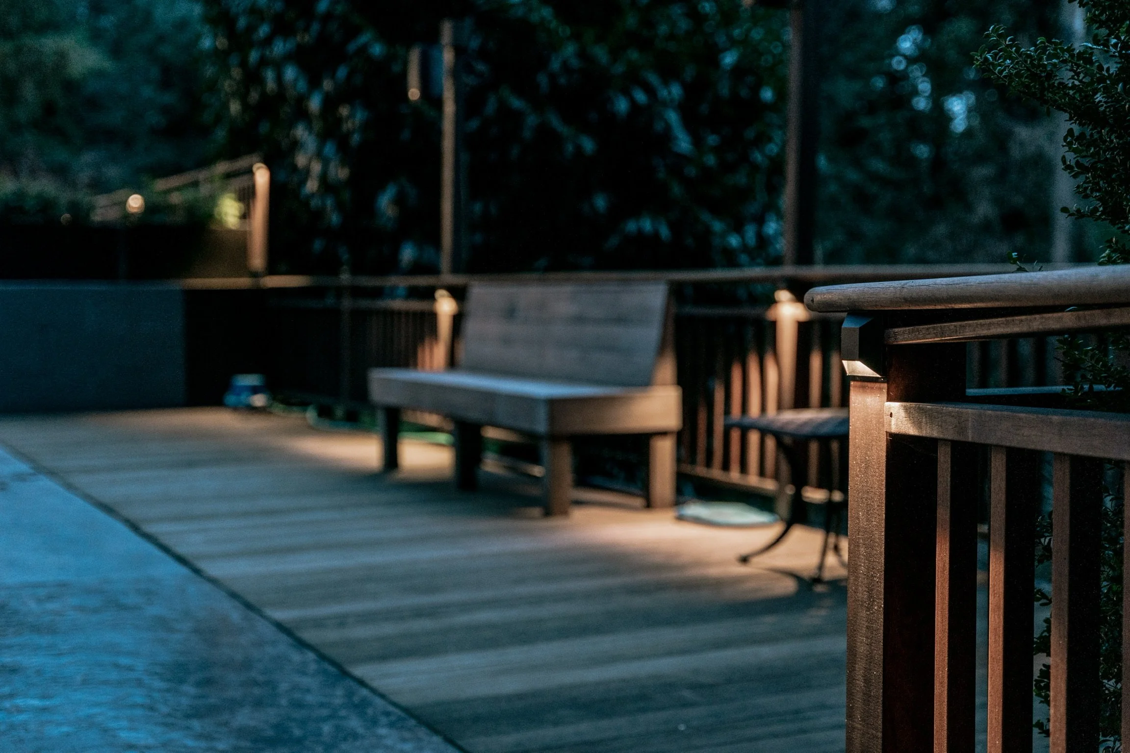 Empty wooden bench on a balcony or patio, with a small table and additional seating, surrounded by greenery and illuminated by outdoor lighting at dusk.