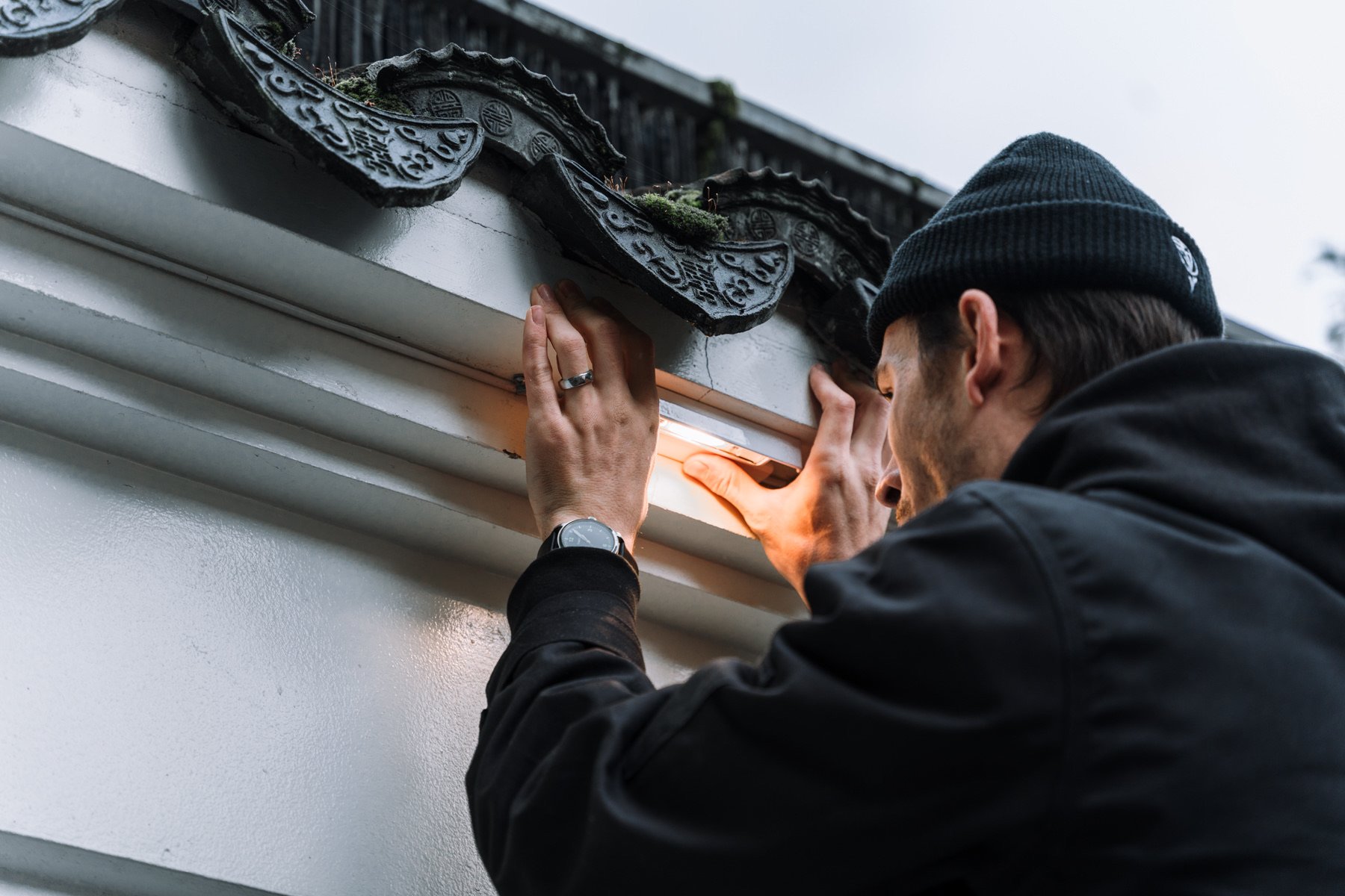A man installing or repairing a light fixture on a white wall outdoors, wearing a black jacket, black beanie, and watch.