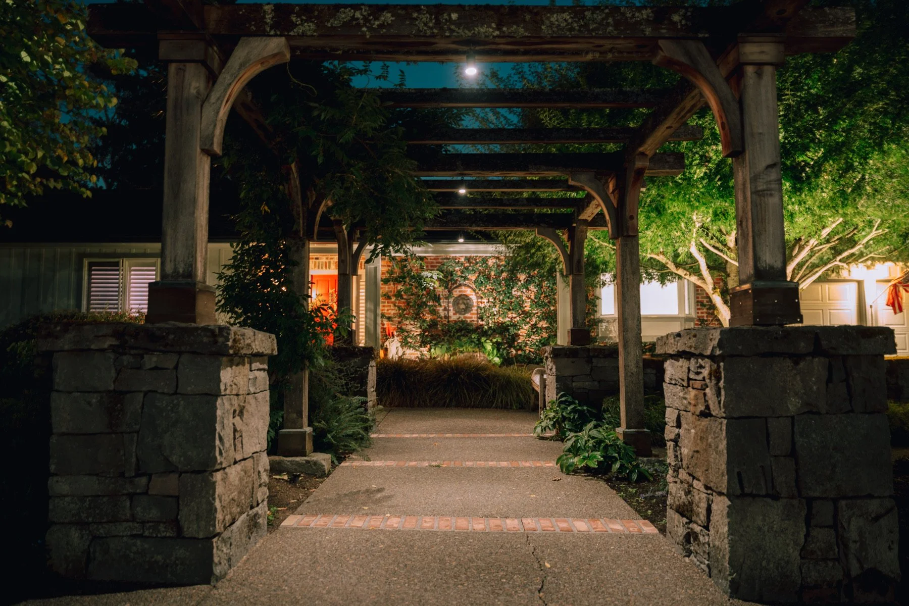 Nighttime view of a wooden garden archway with brick and stone pillars framing a sidewalk leading to a house with illuminated windows and greenery, including trees and shrubs.