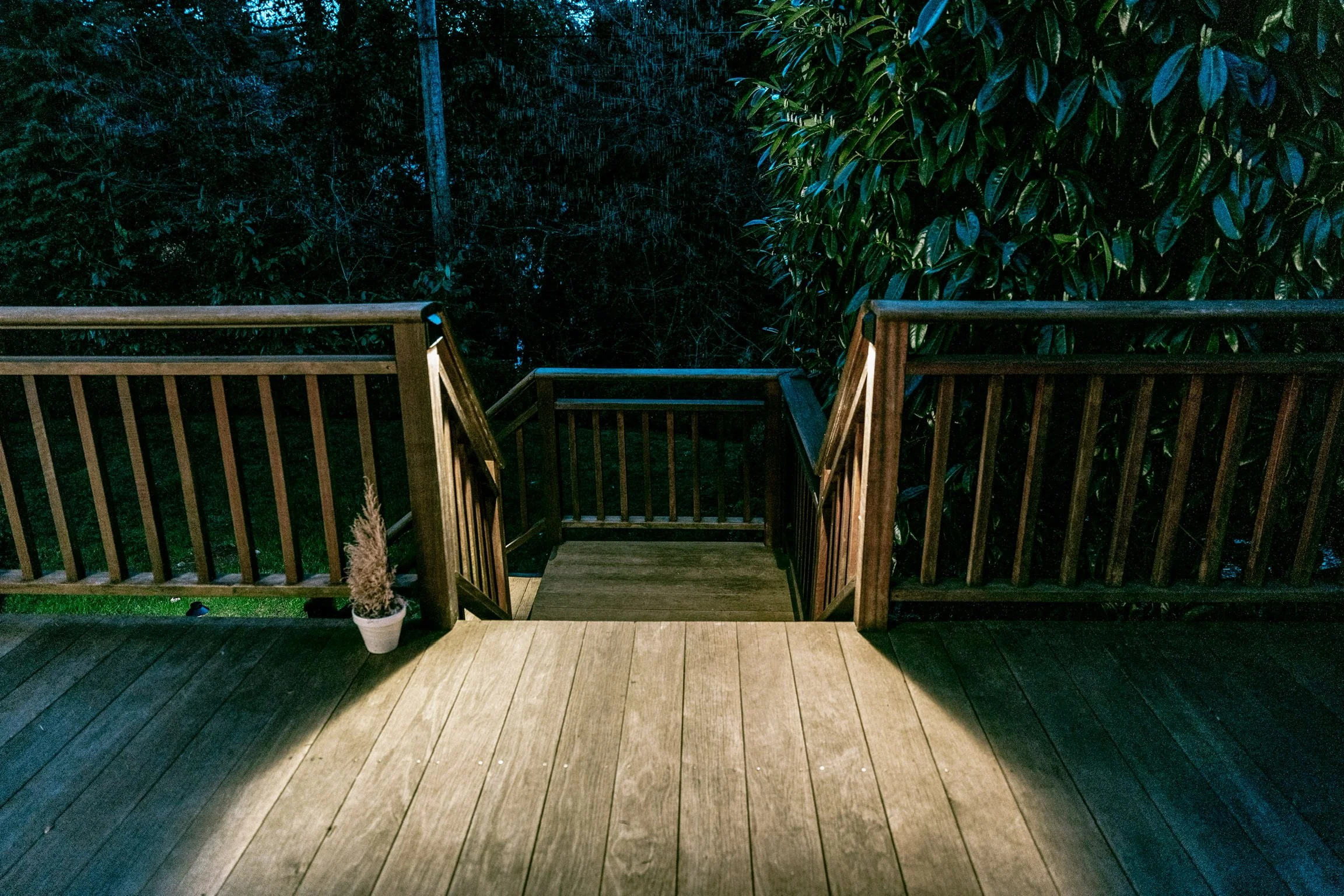 A wooden deck with a railing and stairs leading down to a garden area at dusk or night. There is a potted plant on the deck to the left.