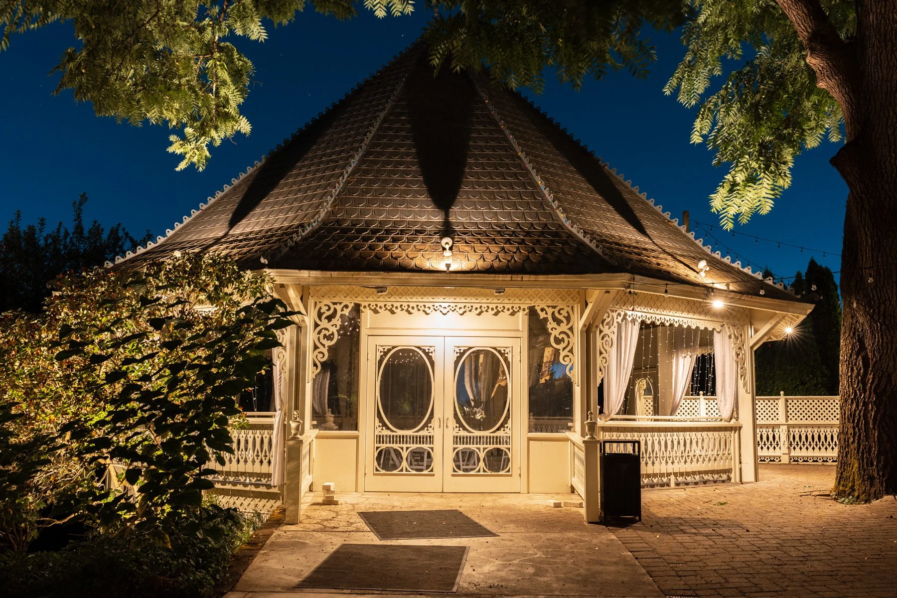 A lit Victorian-style gazebo at night with white ornate woodwork, curtains, and a shingled roof, surrounded by trees and bushes.