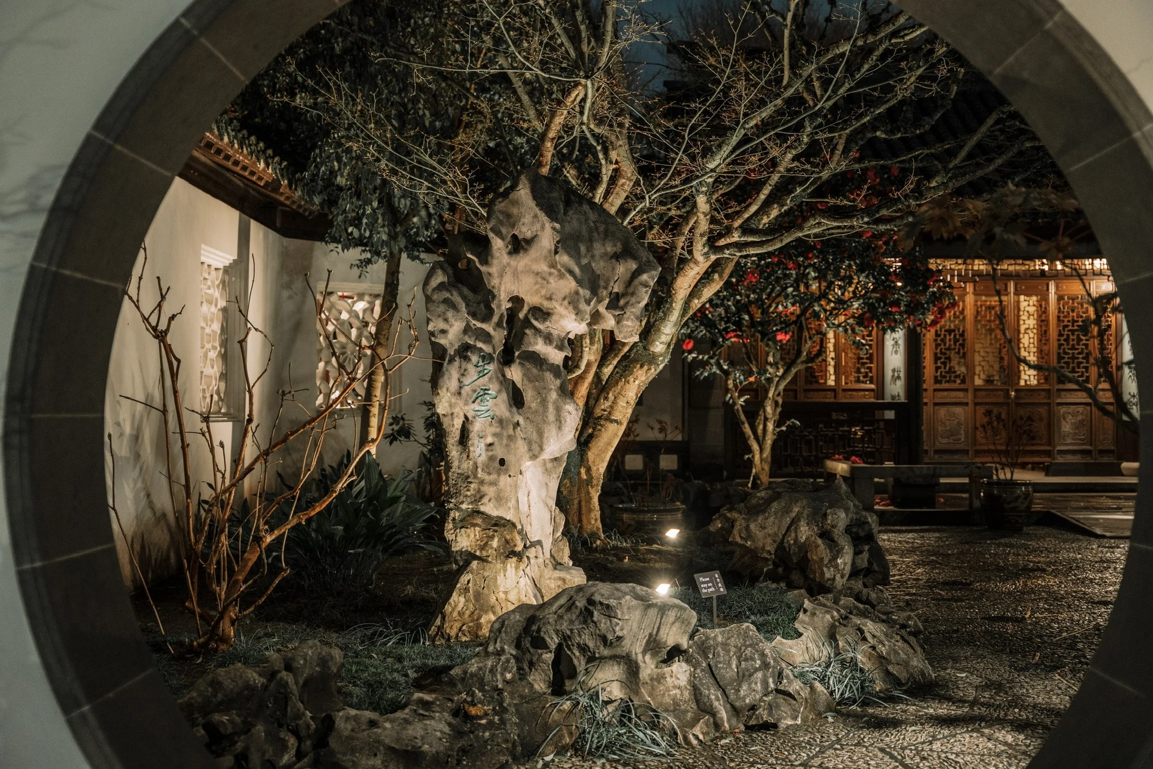 An indoor garden scene viewed through a circular opening, featuring rocks, small plants, a large stone with inscriptions, a tree with red berries, and an intricately carved wooden wall in the background.
