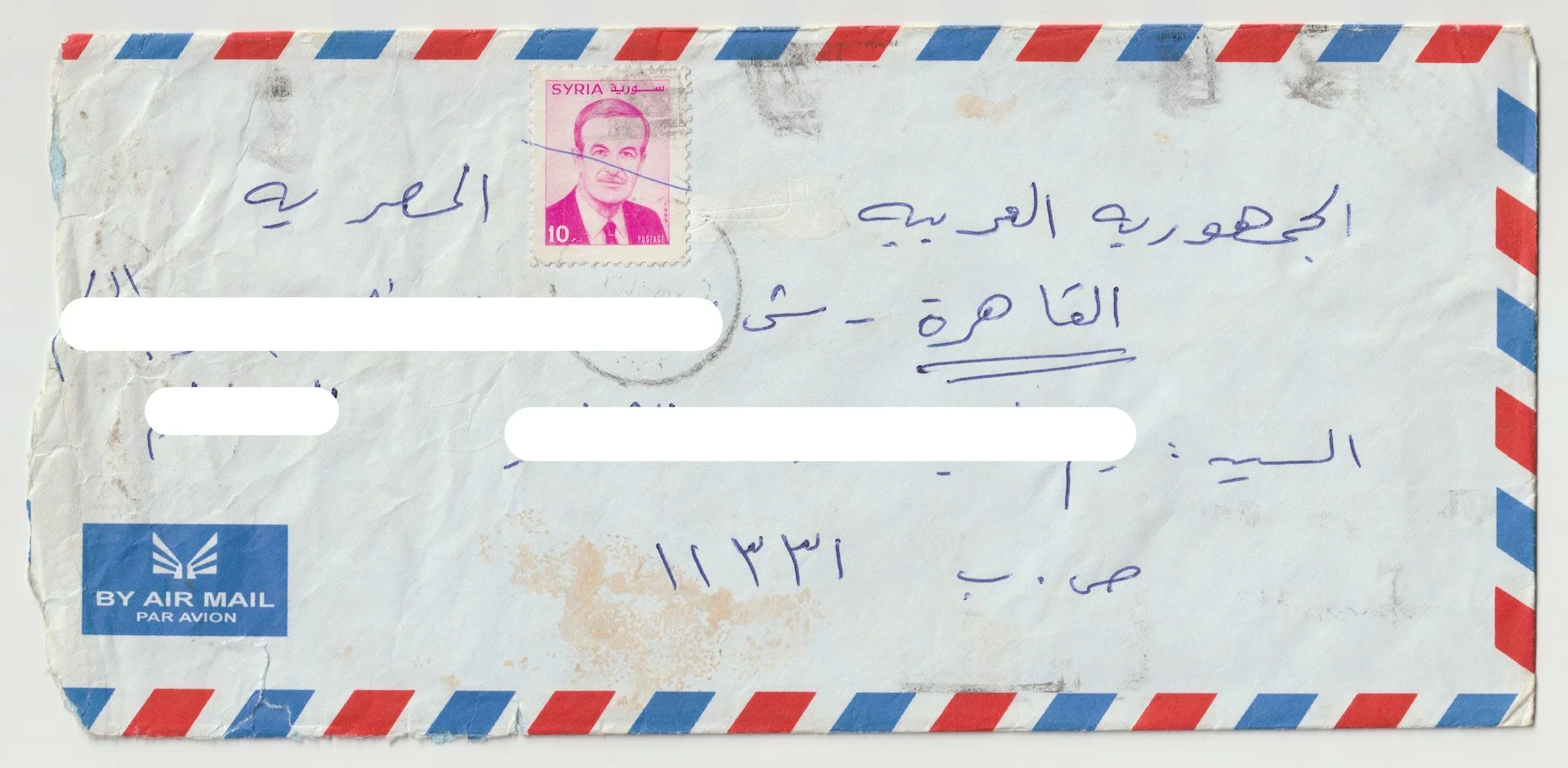 A worn rotary air mail envelope with red and blue striped border and a pink Syria postage stamp featuring a portrait of a man. Handwritten text in Arabic is visible on the envelope.