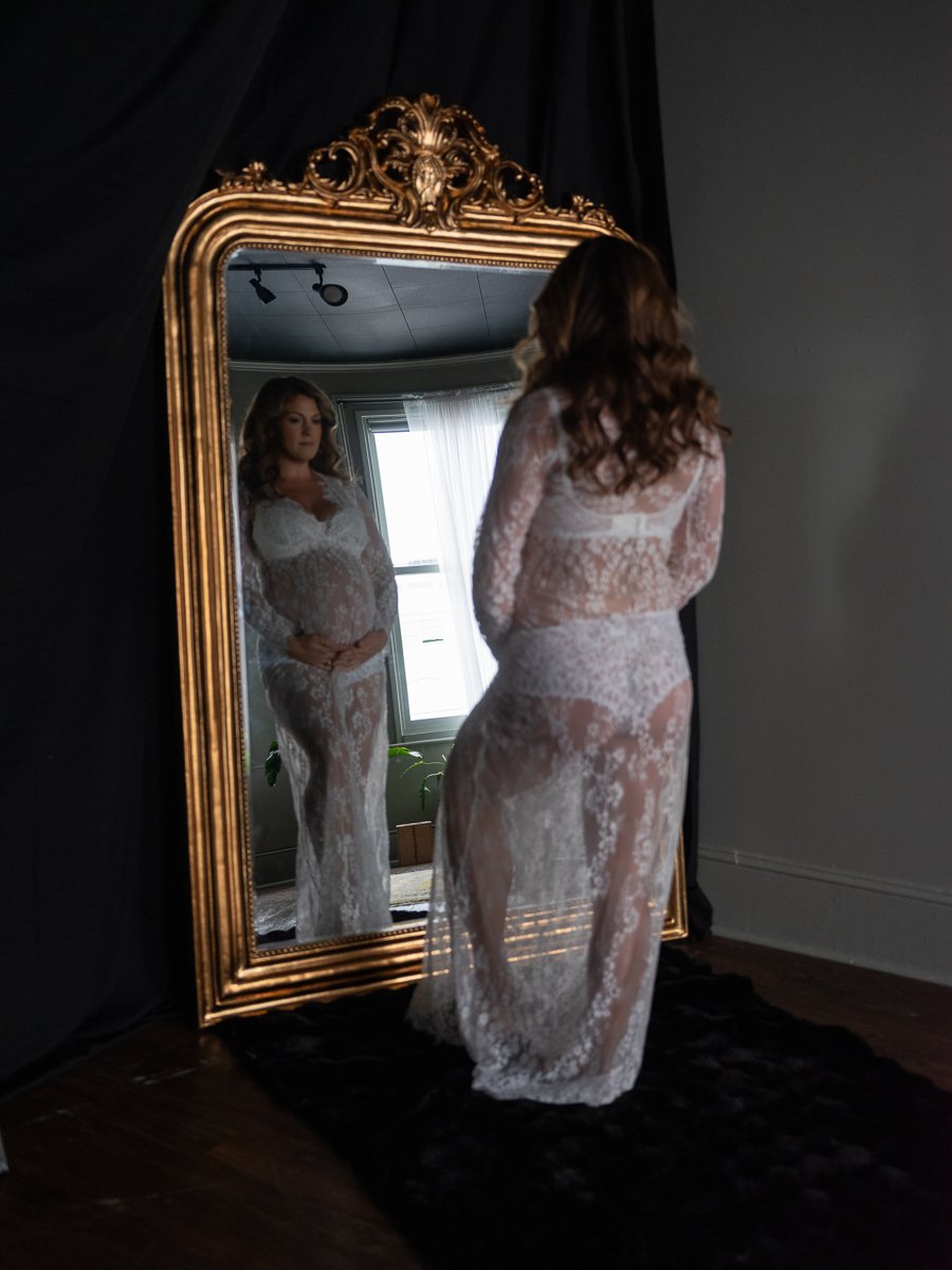 An elegant maternity boudoir portrait of a pregnant woman in a sheer lace gown reflected in an ornate gold mirror, photographed at Bay Street Studios, Beaufort, SC.