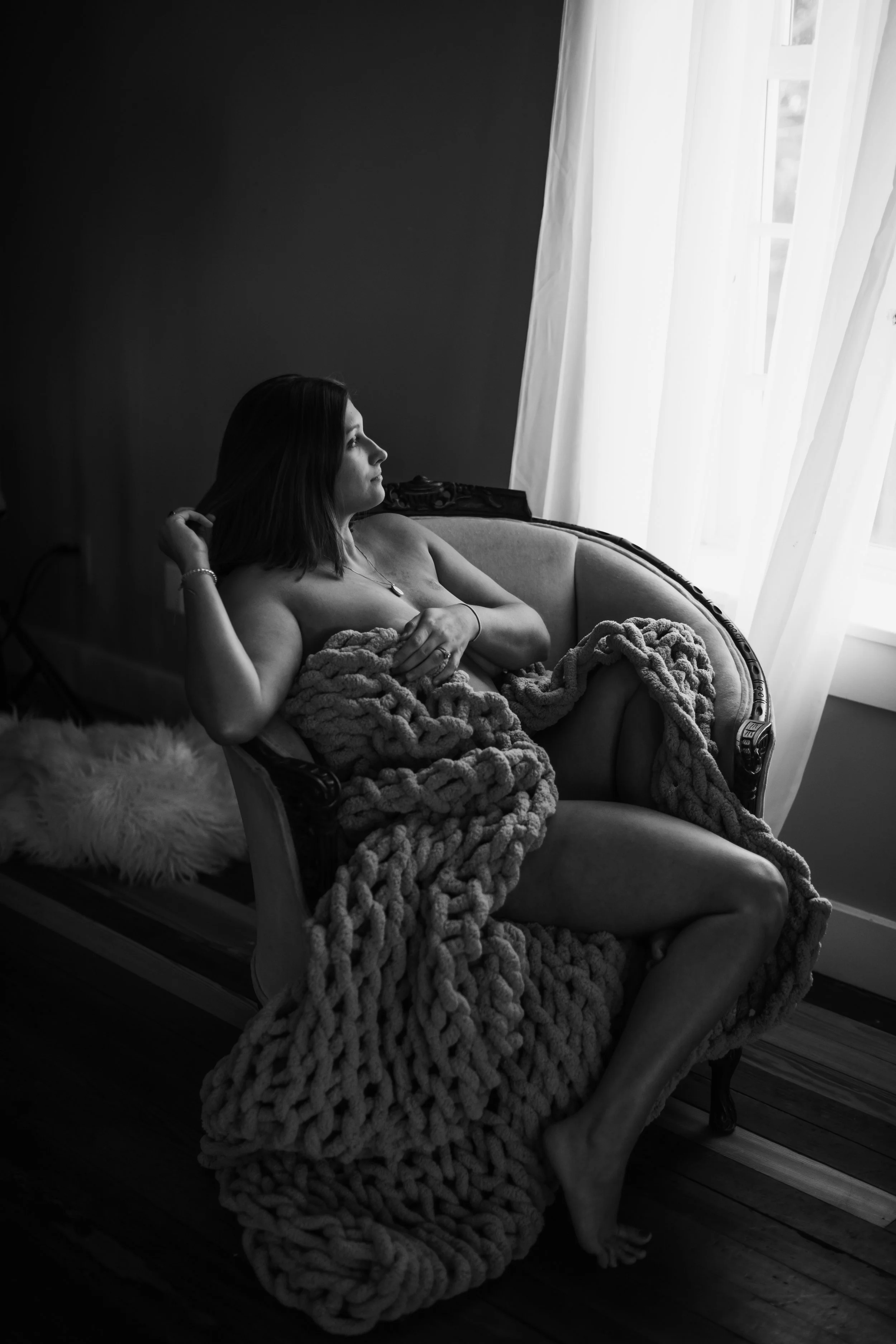 Woman reclining in a vintage armchair wrapped in a chunky knit blanket, soft natural light raw portrait by Emily Scott