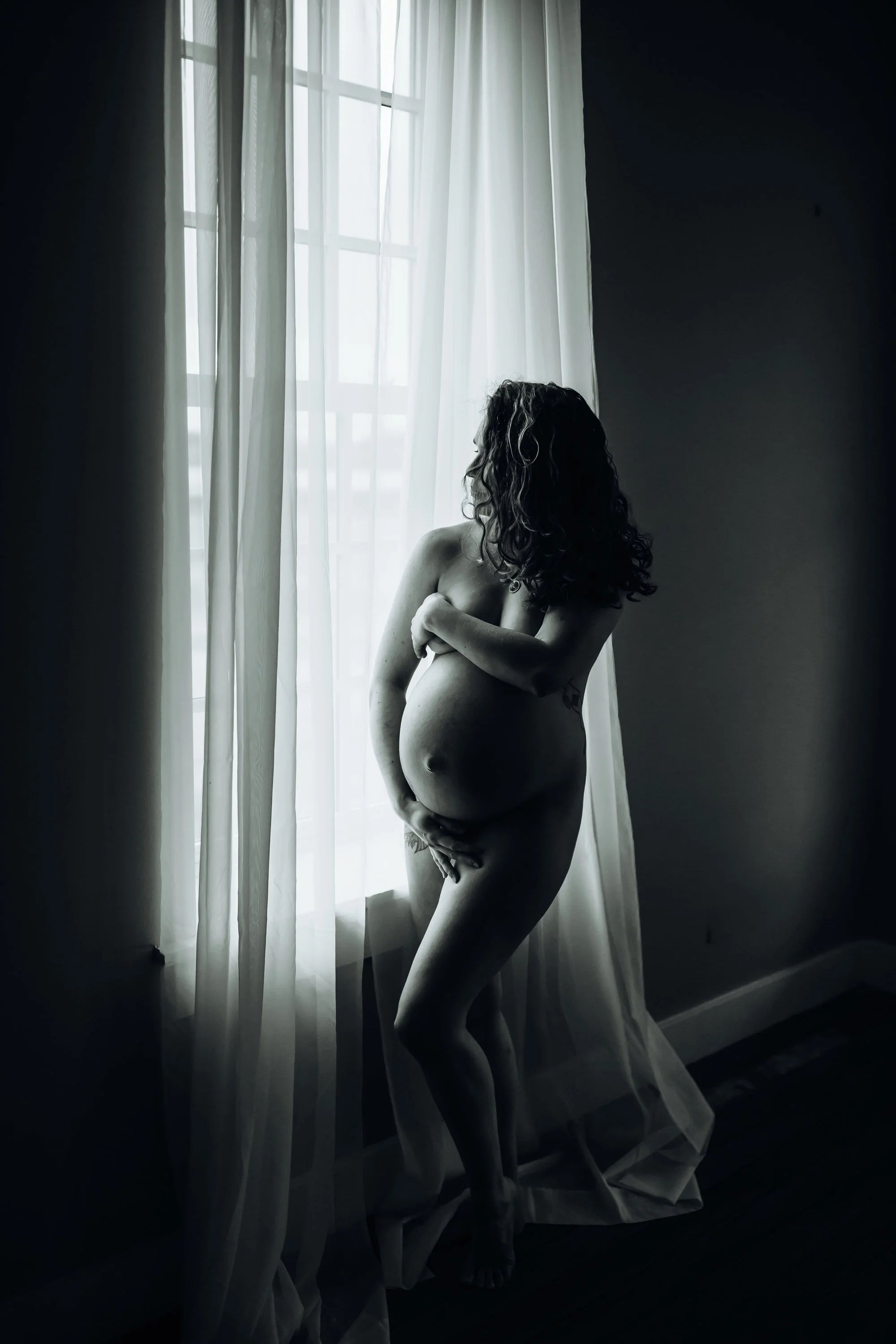 A stunning black and white fine art maternity boudoir portrait of a pregnant woman cradling her belly in dramatic natural window light, photographed by Emily Scott in Beaufort, South Carolina.