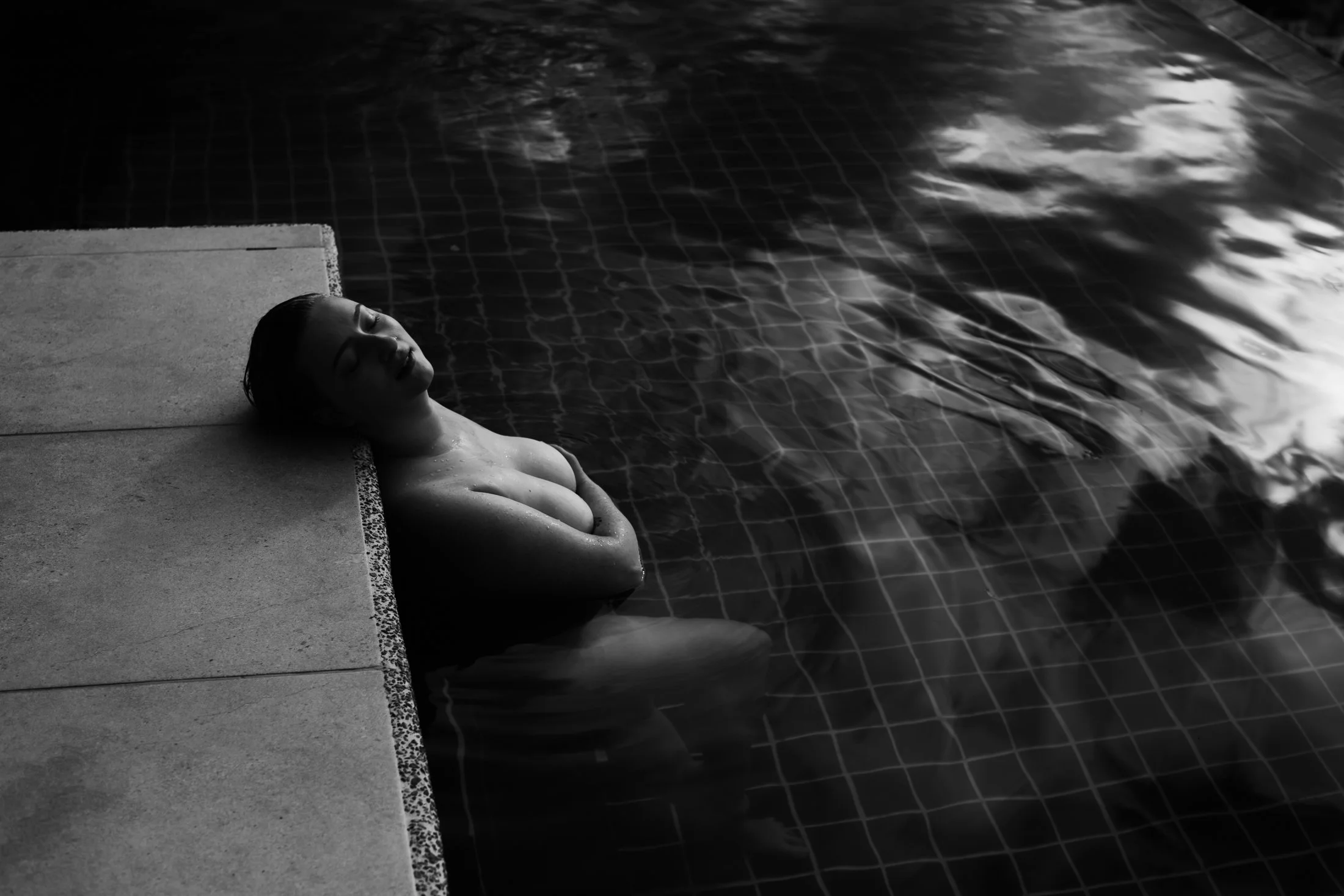 A black and white fine art portrait of a woman floating at the edge of a pool with dramatic water reflections, photographed as part of The Raw Project by Emily Scott in Beaufort, SC.
