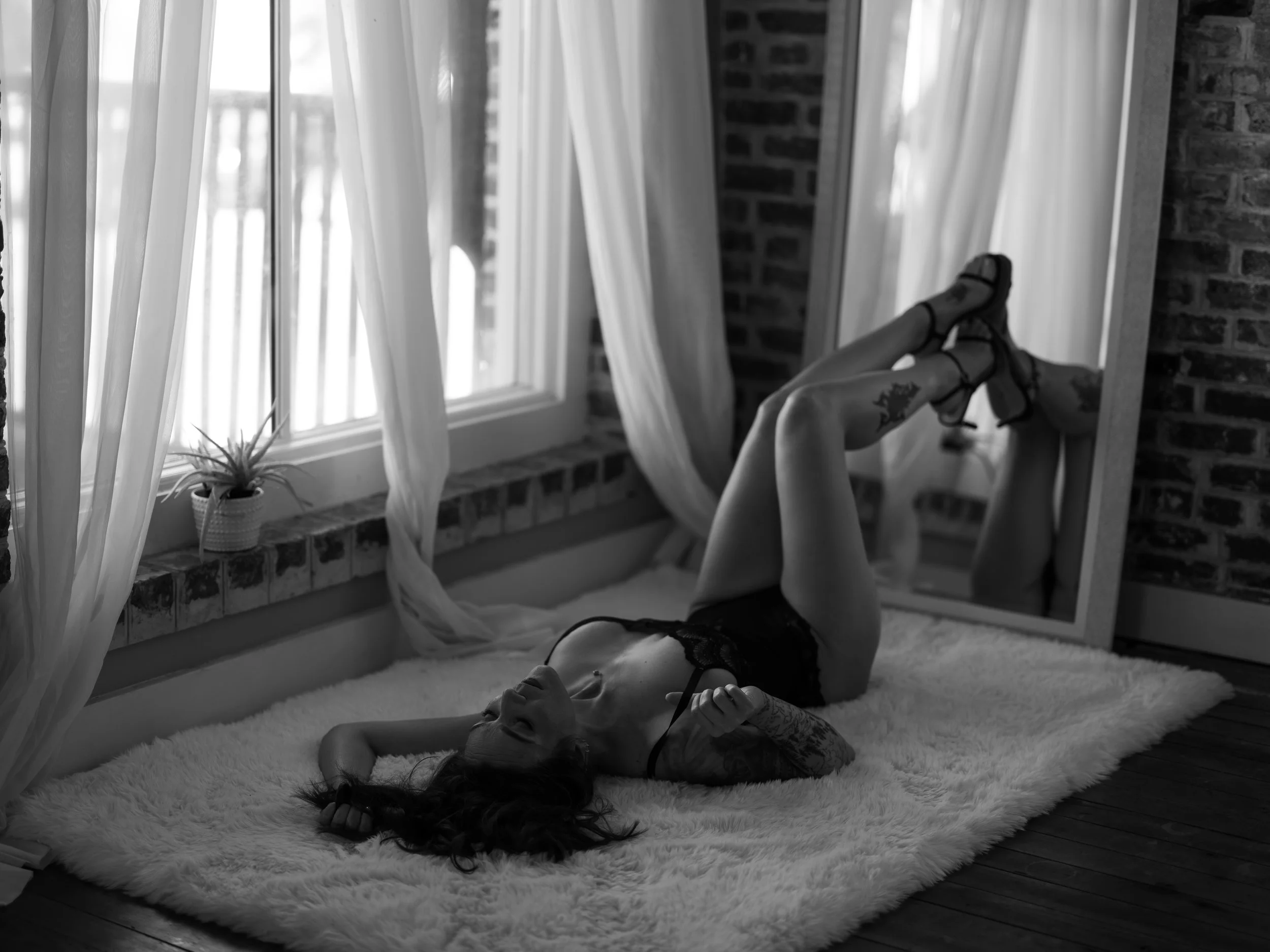 A woman with tattoos lying on a fluffy white rug, wearing black lingerie and sandals, with legs raised and reflected in a mirror. Natural light coming through a window with sheer curtains, and a potted plant on the windowsill.