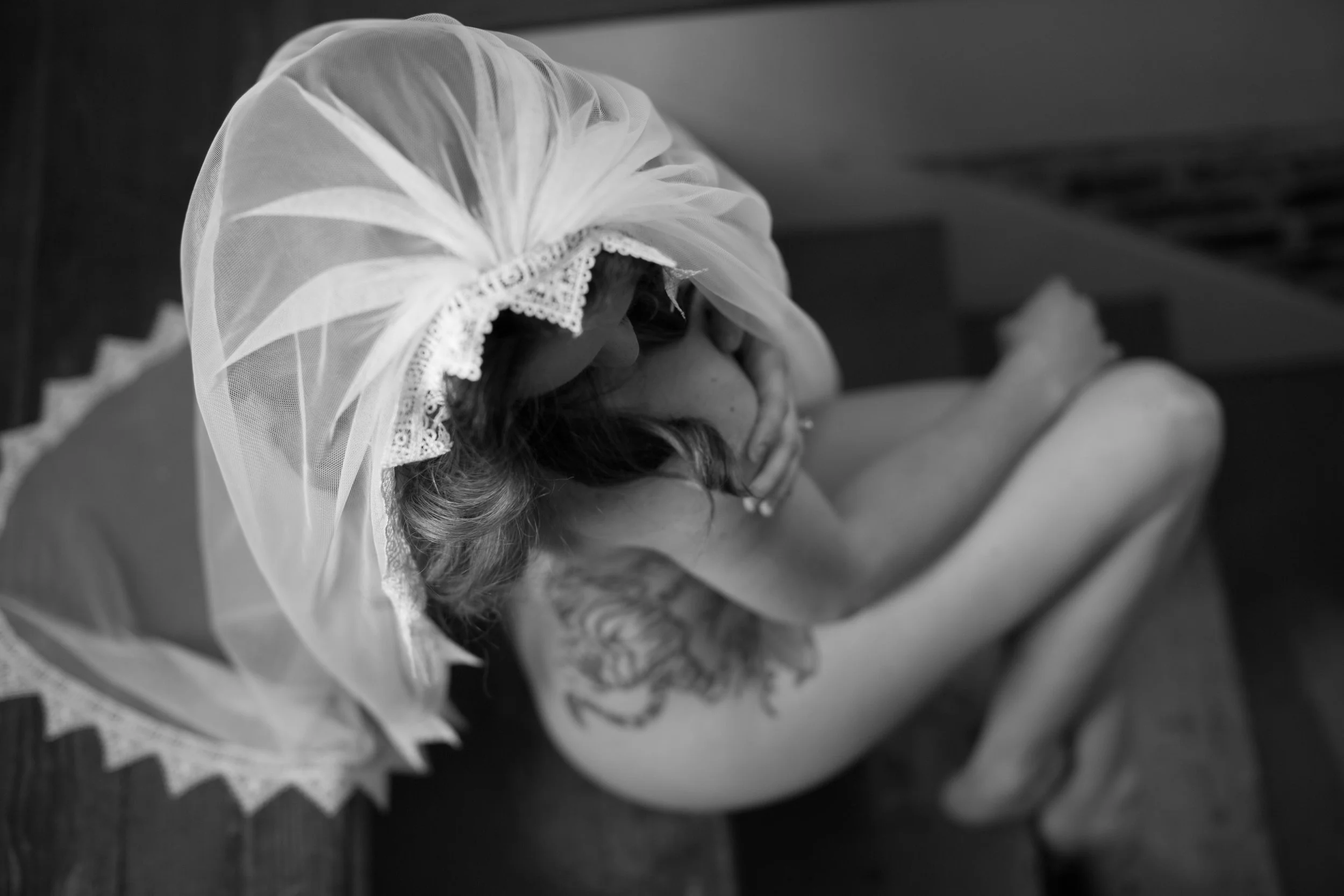 A woman with a tattoo on her shoulder sitting on a bench, wearing a lace veil over her head, covering her face and eyes, in black and white.