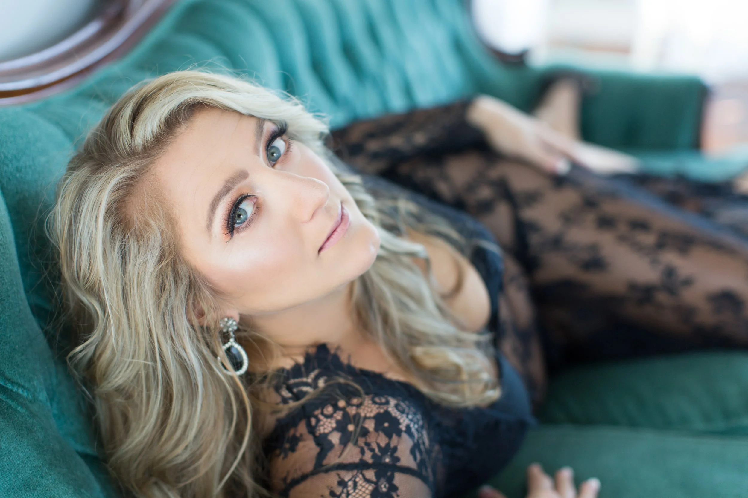 A woman with blonde hair and blue eyes lies on a green velvet couch, dressed in black lace attire and earrings.