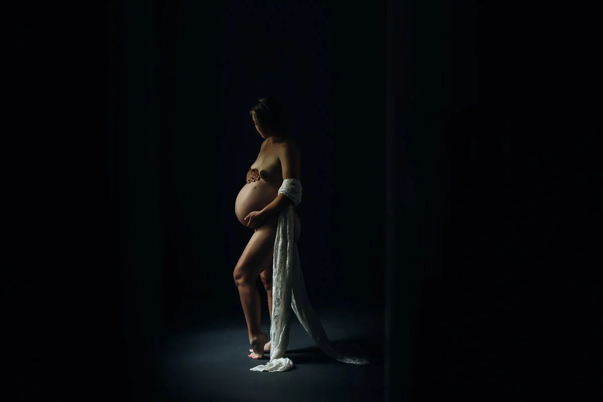  An intimate maternity boudoir portrait of a pregnant woman cradling her belly in soft dramatic light, photographed by Emily Scott in Beaufort, SC.