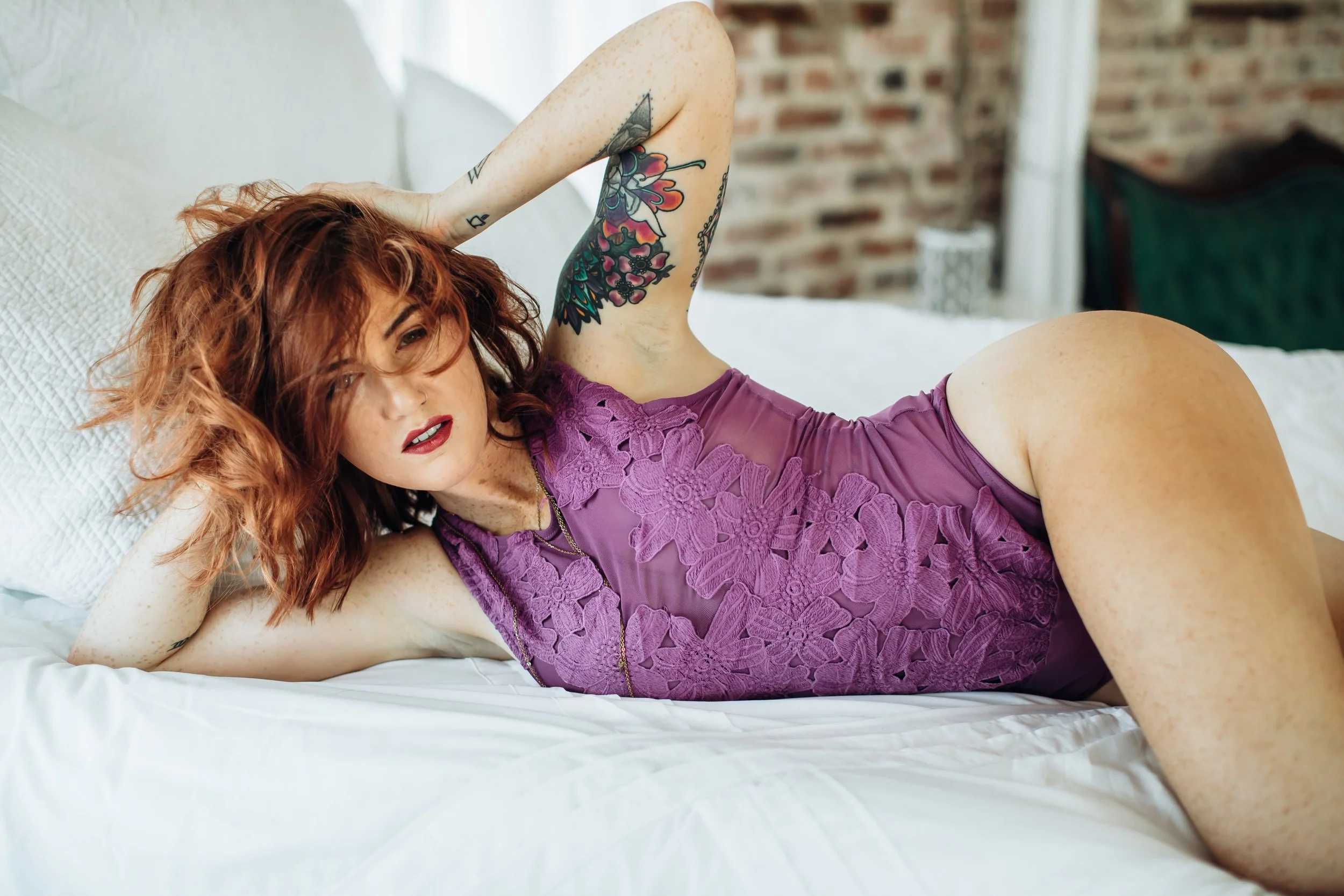 A woman with red hair and tattoos lying on a bed, wearing a purple lace dress.