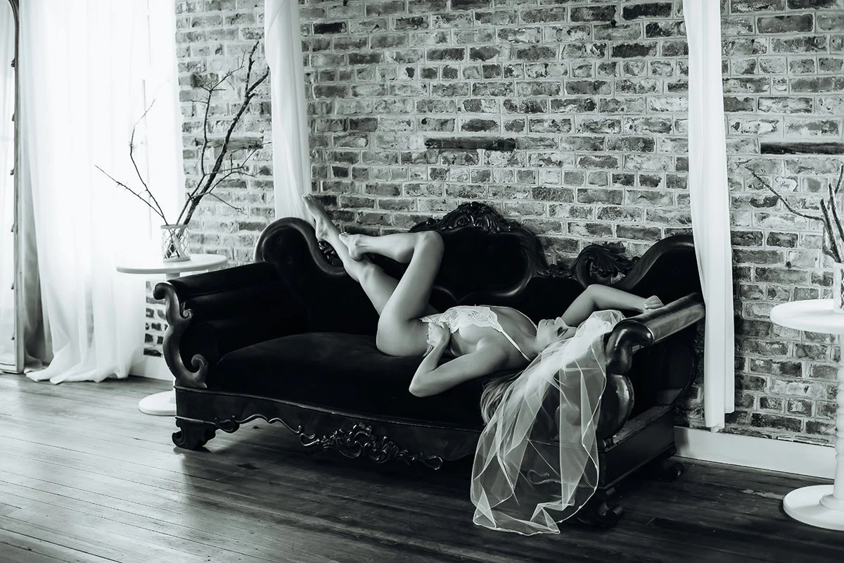  A dramatic black and white bridal boudoir portrait of a bride reclining on an antique velvet chaise with a flowing veil, photographed by Emily Scott in Beaufort, SC.